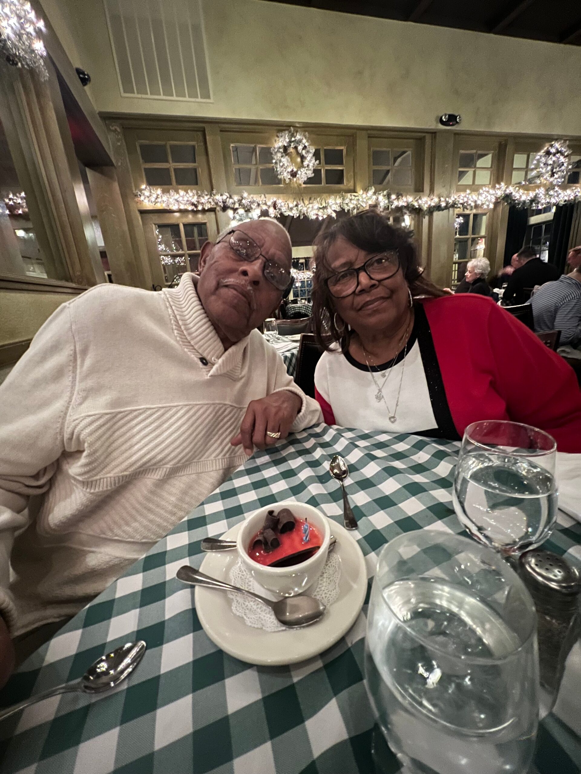 Two smiling seniors sit at a green-and-white checkered table, with dessert and water glasses in front, festive lights in the background.
