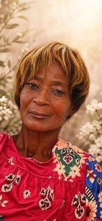 Portrait of an older woman with short light brown hair wearing a colorful floral blouse, standing in a sunlit garden with white flowers in the background.