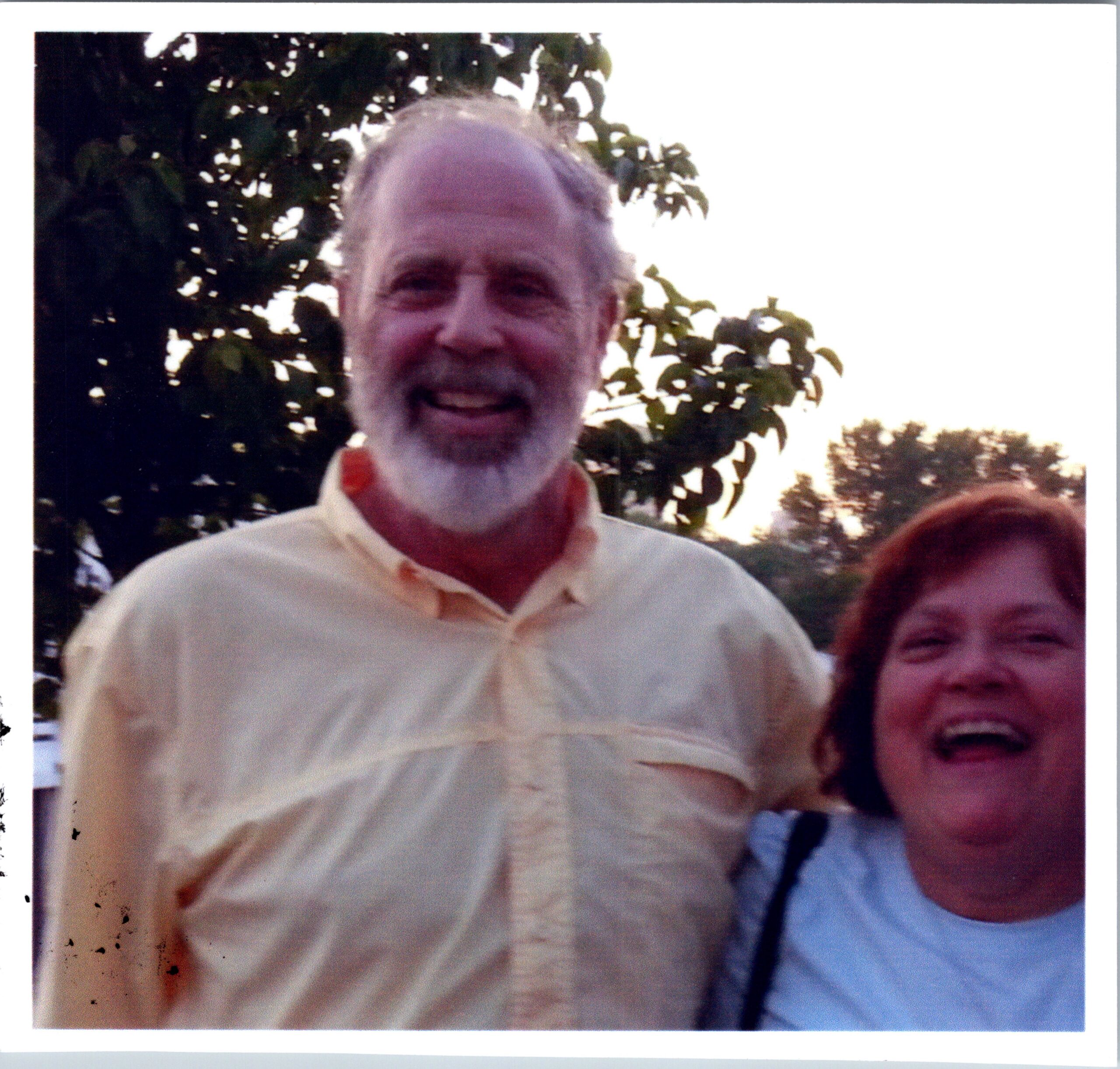Smiling older man with gray beard outdoors, wearing a light shirt, with trees in the background; another person partially visible on the right.
