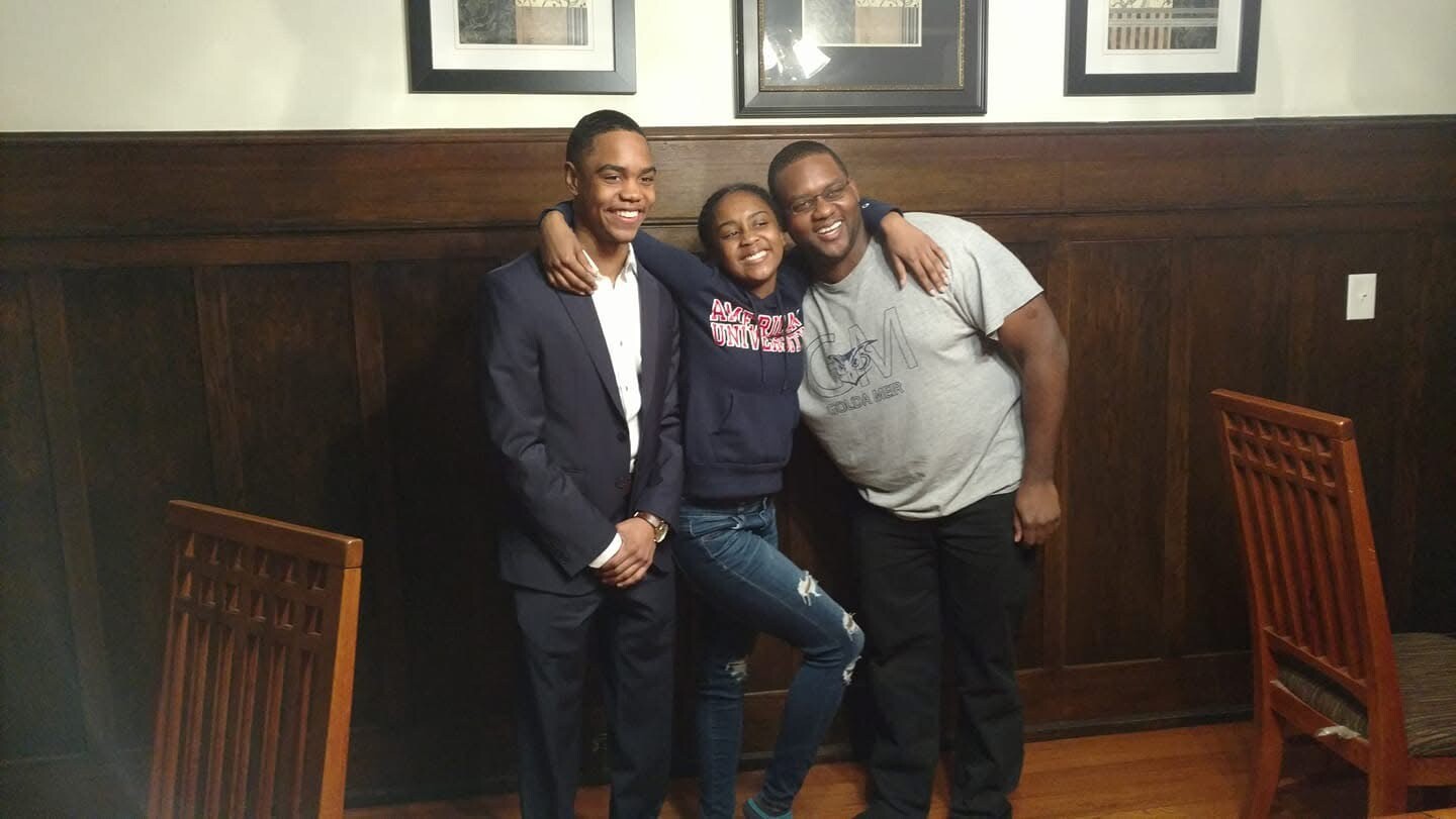 Three friends pose together indoors, smiling with their arms around each other in front of a dark wood-paneled wall with framed pictures above.