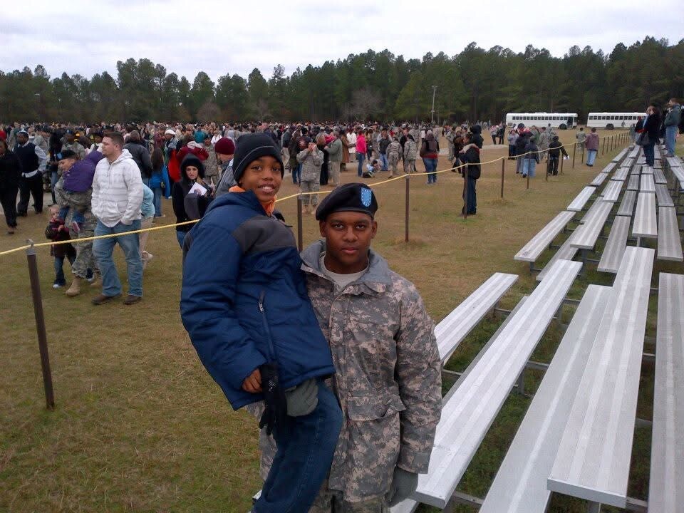 Two people posing near metal bleachers at an outdoor event with a large crowd and buses in the background.