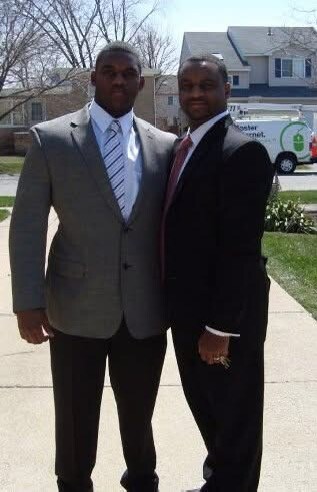 Two men in suits posing together on a sunny suburban sidewalk, with houses and a van in the background.
