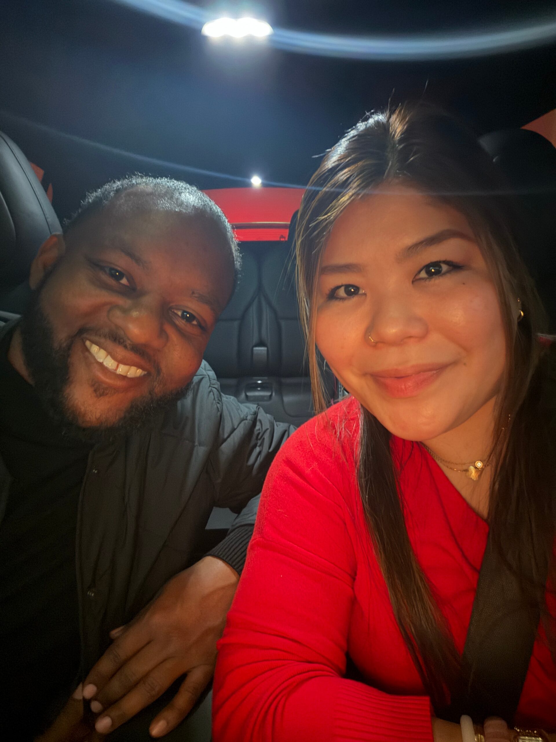 Two friends taking a close-up selfie in a car at night; both are smiling, with the woman in a red top wearing a seatbelt on the right and the man on the left in a dark jacket.