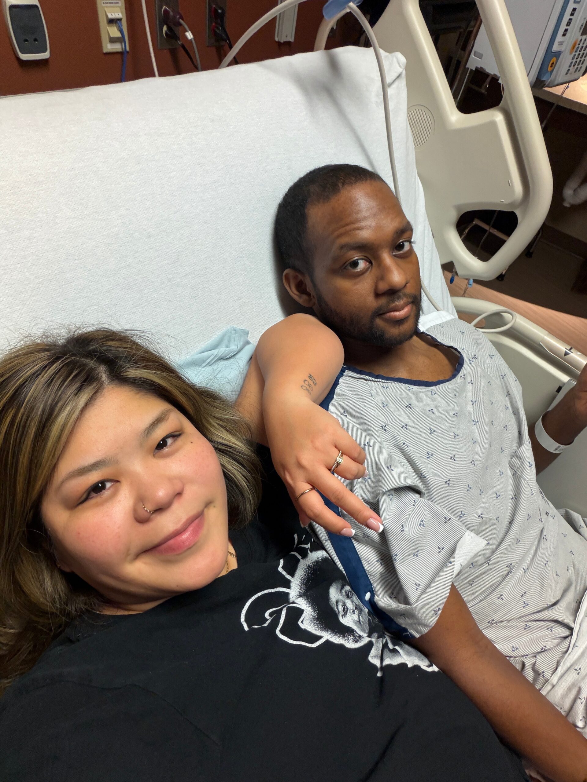Two people nurse a selfie in a hospital room: a man in a patient gown sits up in bed while a woman beside him smiles at the camera, medical equipment in the background.