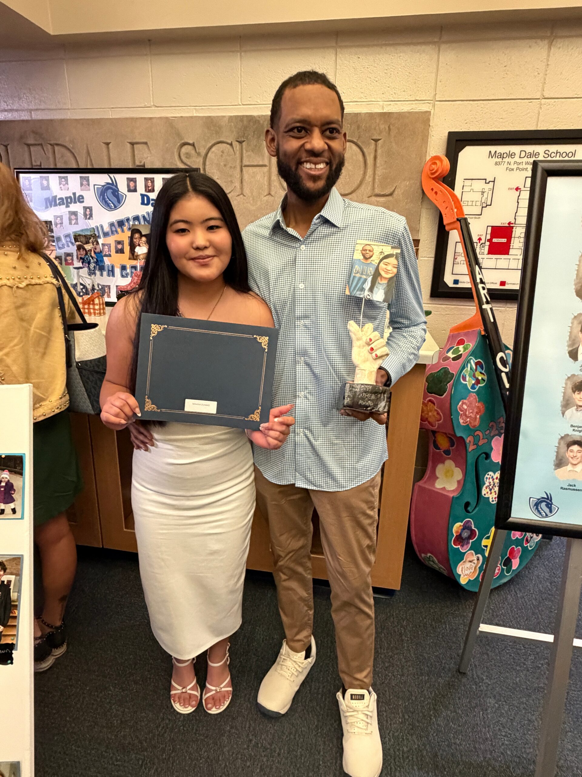 Two students pose together in a school hallway; the girl holds a diploma and the guy holds a trophy and a photo, smiling at the camera.