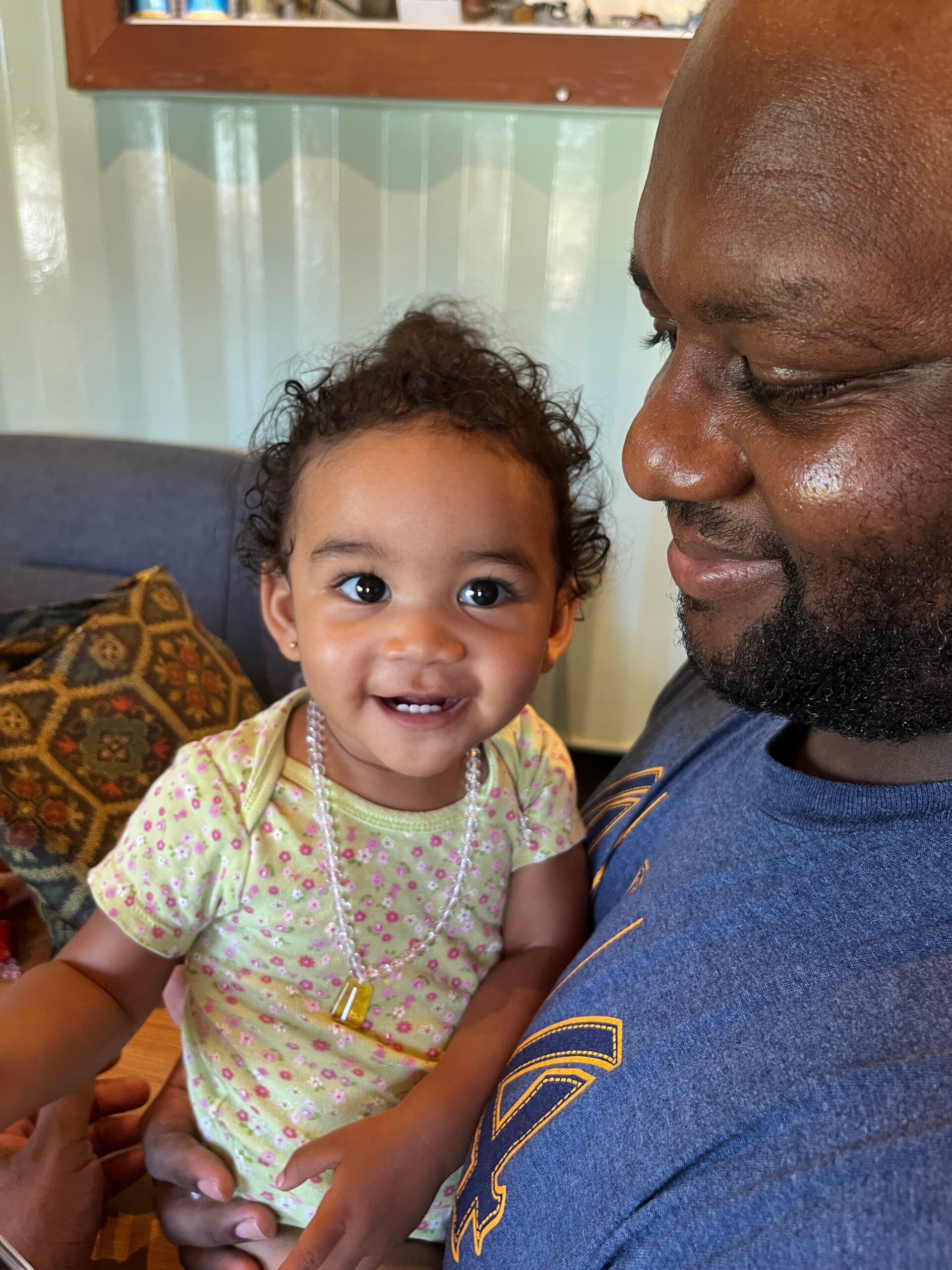 Adult man holding a smiling baby girl indoors, faces close together and looking at each other.