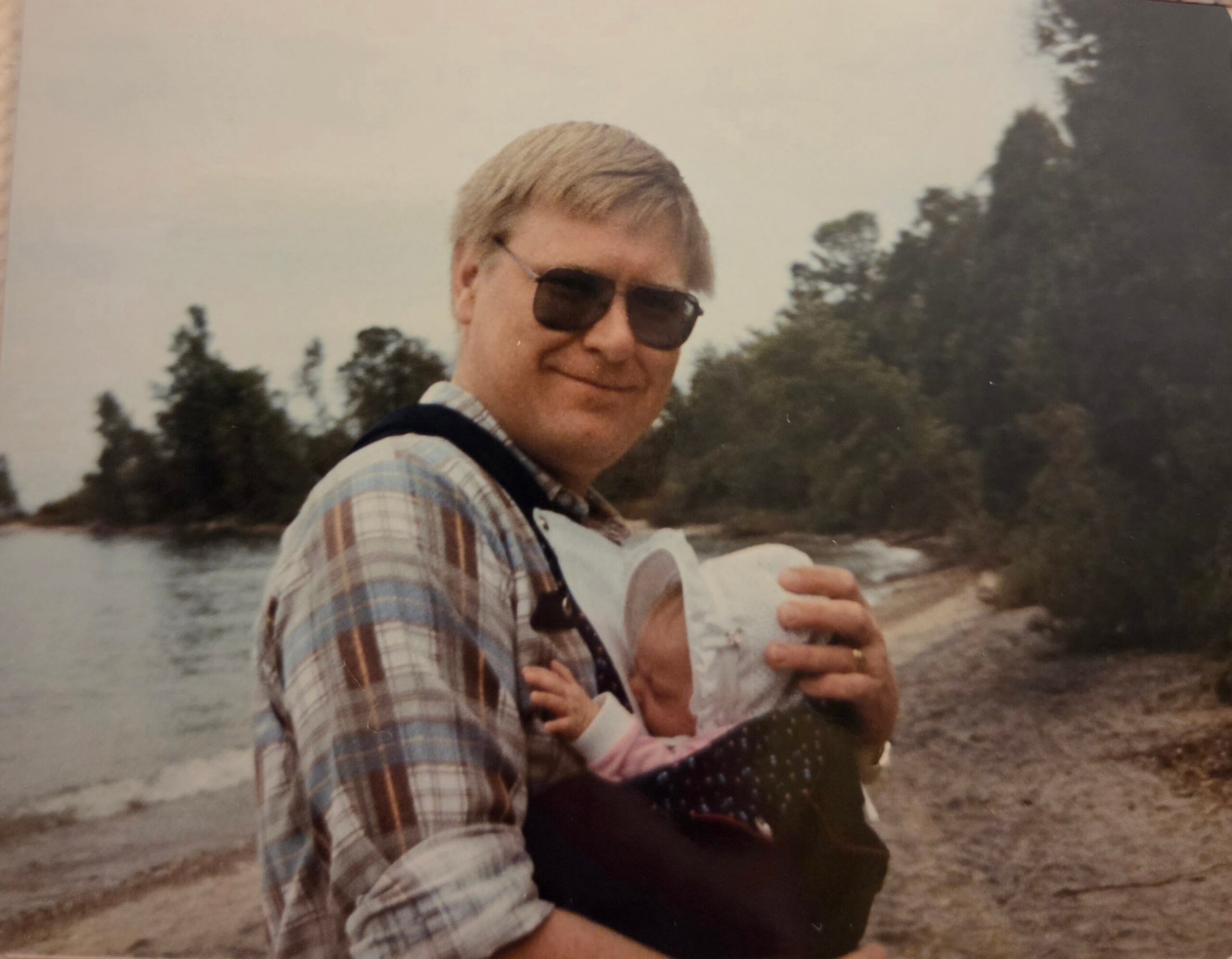 Man wearing sunglasses and a plaid shirt holds a baby wrapped in a blanket at a lakeside/beach area with trees in the background.