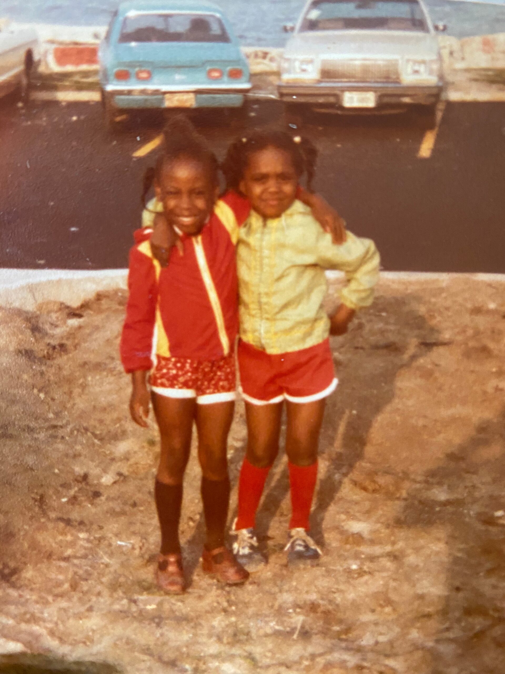 Two young girls stand side by side with arms around each other in a parking lot, wearing bright red and yellow outfits, vintage cars in the background.