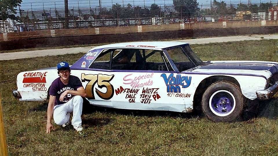 Vintage stock car with number 75 parked on a grassy infield; man in a cap kneels beside the car.
