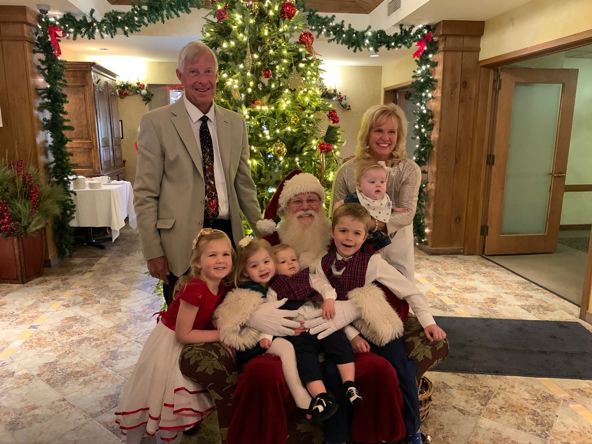 Family posing with Santa Claus in a festive lobby scene with a large Christmas tree and garlands behind them.
