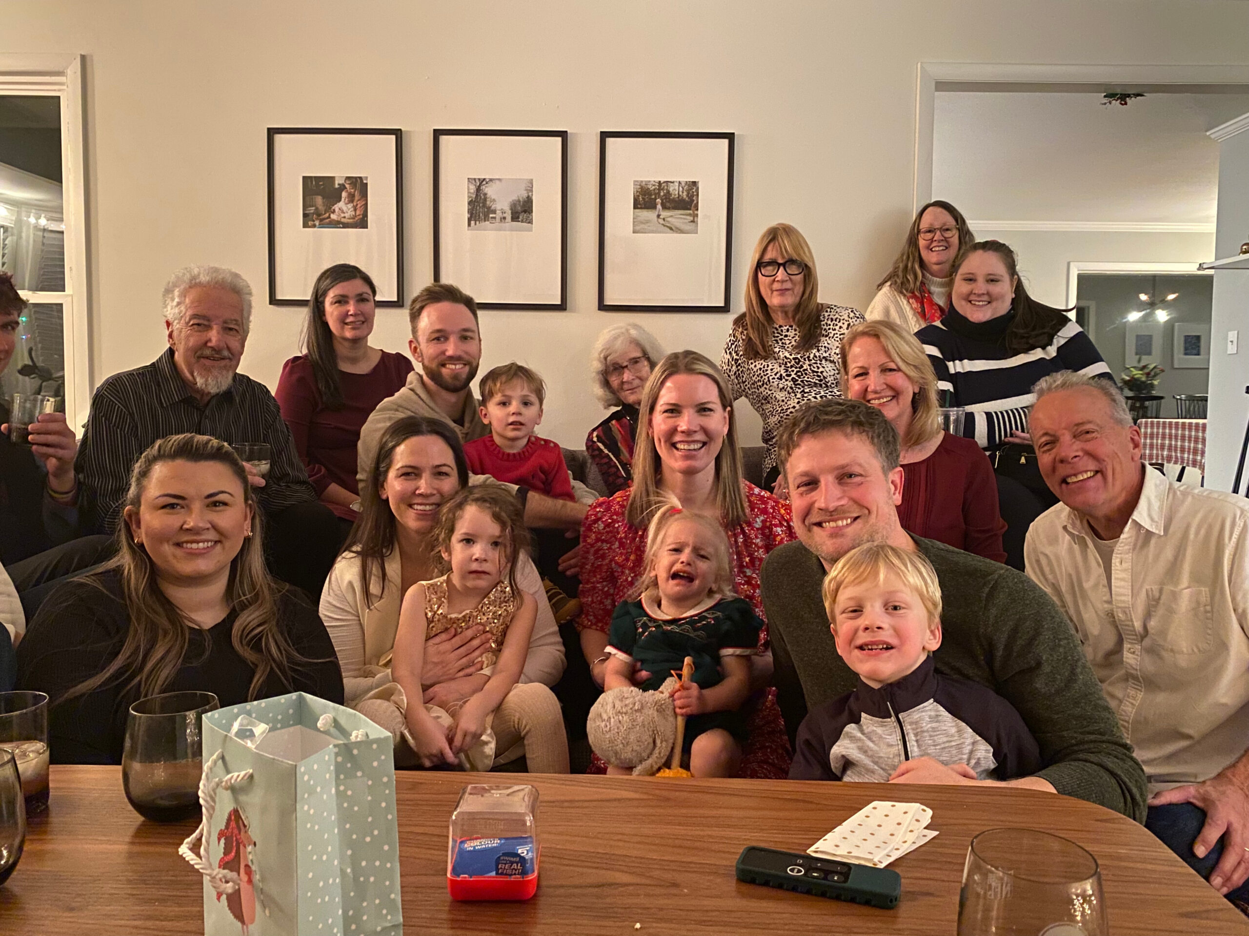 Group of smiling adults and children posing for a festive photo around a dining table with gift bags in the foreground and framed pictures on the wall behind them.