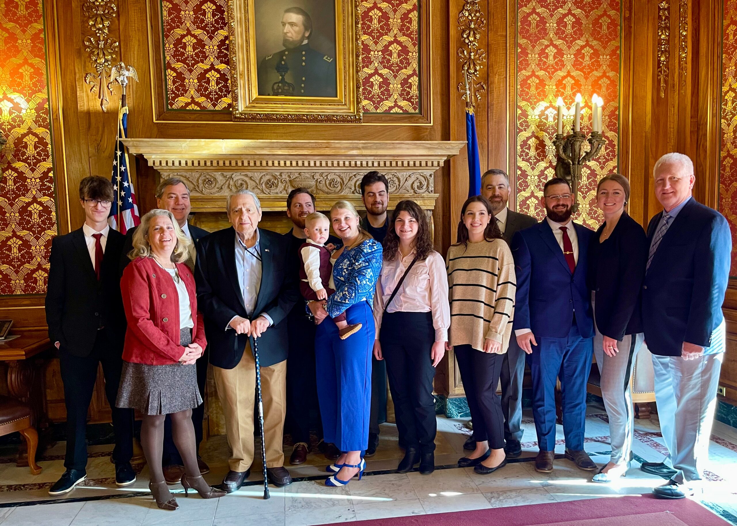 Group of people in formal attire posing in a richly decorated room with a portrait above a marble fireplace and a U.S. flag nearby, suggesting a formal gathering.