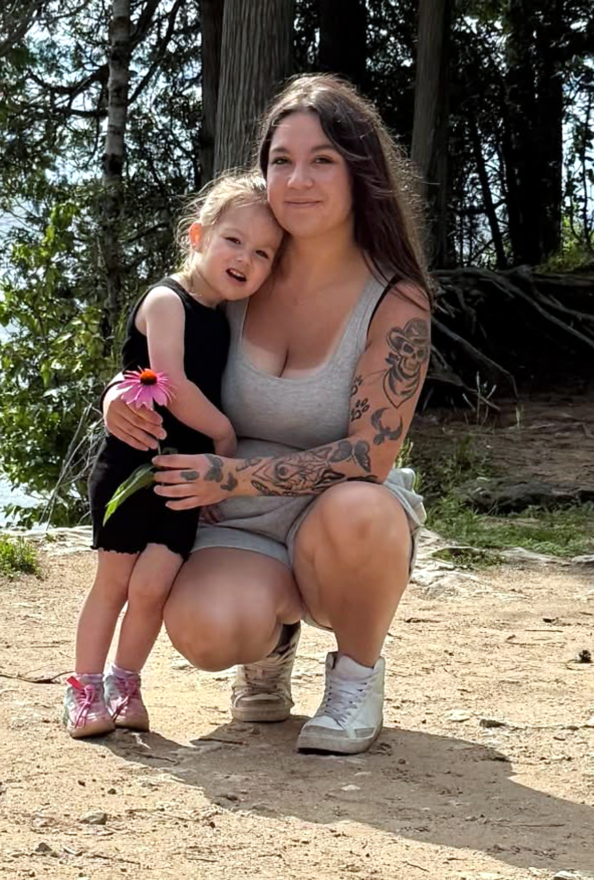 Mother crouching on a dirt path with her arms around a young girl who holds a pink flower, outdoors among trees at the shore.