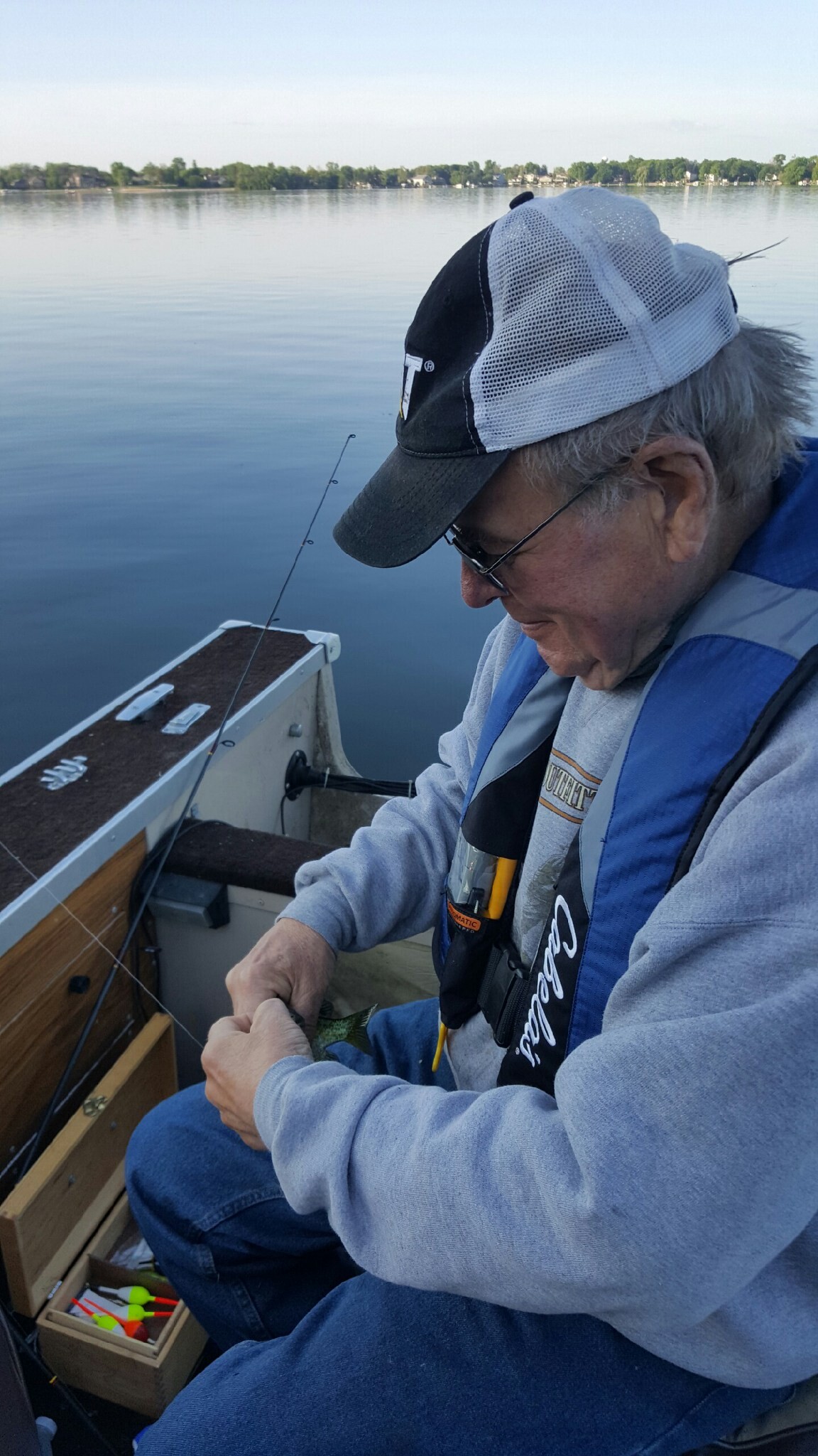 Man in a blue life jacket and cap sits in a small boat on a calm lake, handling a caught fish with a tackle box nearby.