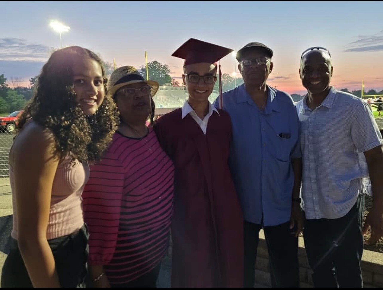 Group of five people posing for a photo at sunset; the graduate wears a maroon cap and gown in the center with family and friends around him/her at a sports field venue.