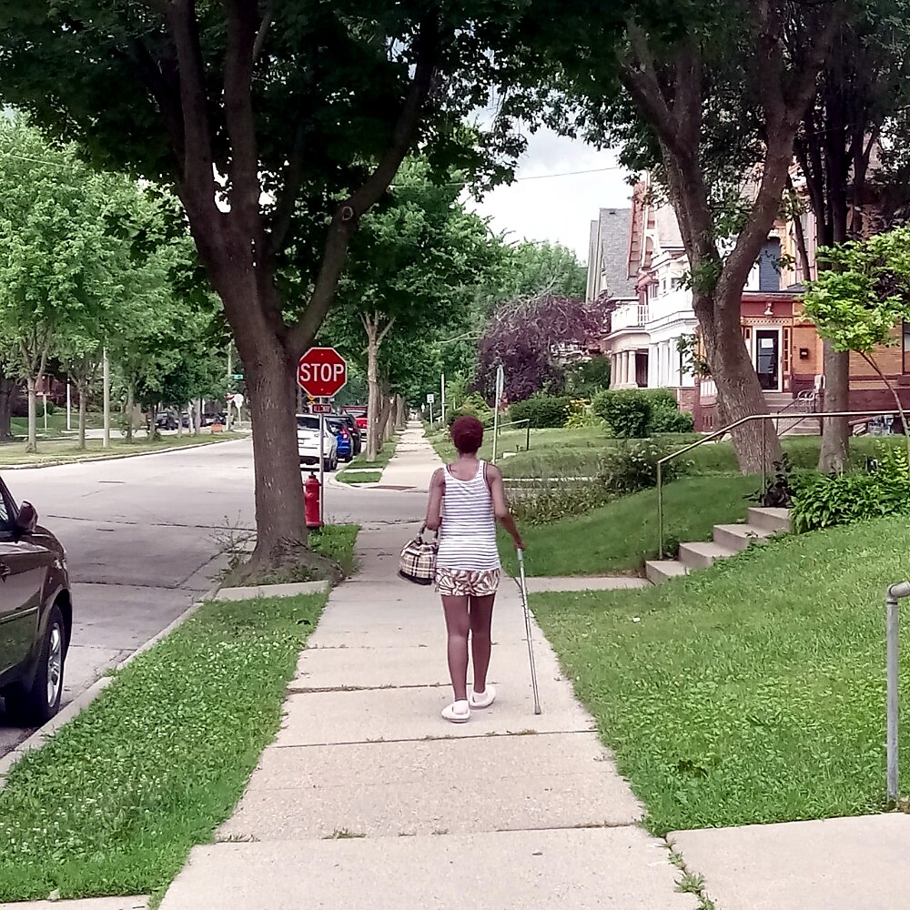 Person with a white striped tank top and shorts walking with a cane on a suburban sidewalk, seen from behind, near a stop sign and green lawns.