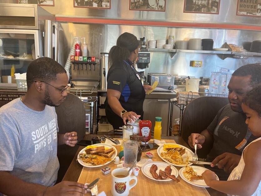 Three diners sit at a wooden table in a casual diner, meals plated before them as a server cleans a coffee area in the back.
