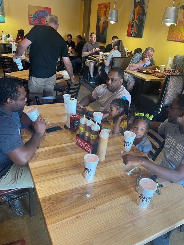 Family and friends dine at a casual restaurant around a long wooden table with drinks; kids are present and condiments sit in the center.