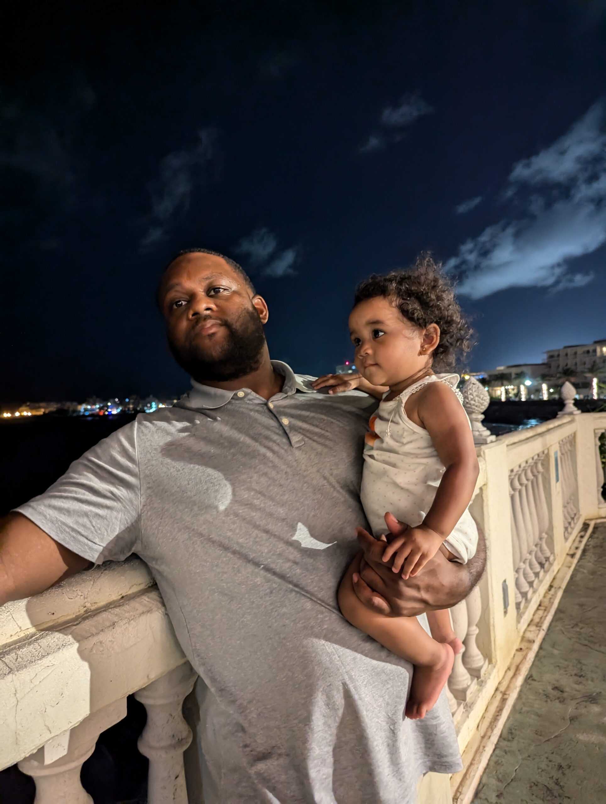 Man holding a young girl on a balcony at night, city lights in the background.