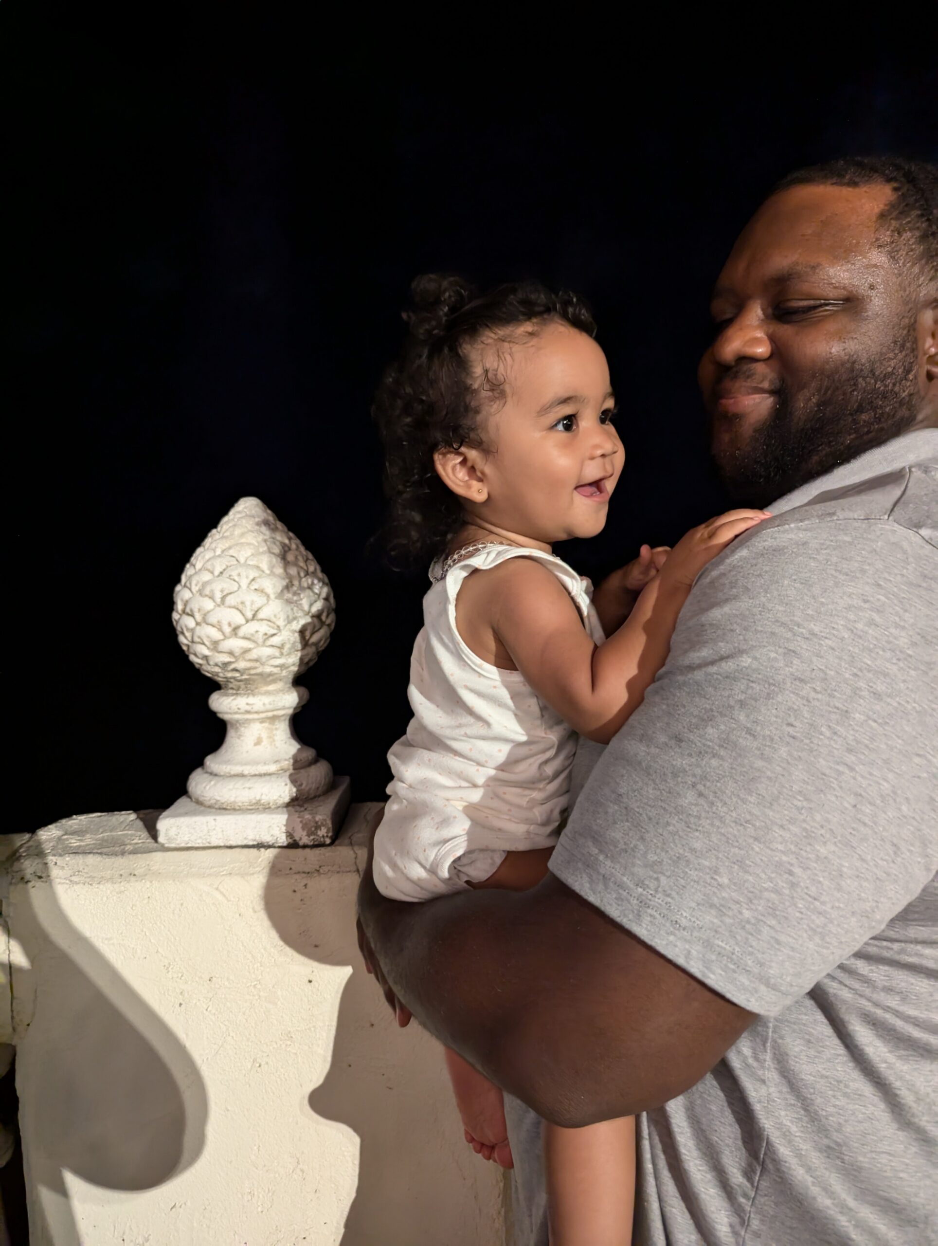 Father in a gray shirt holds a smiling toddler in a white romper on a balcony, with a white decorative post nearby against a dark background.