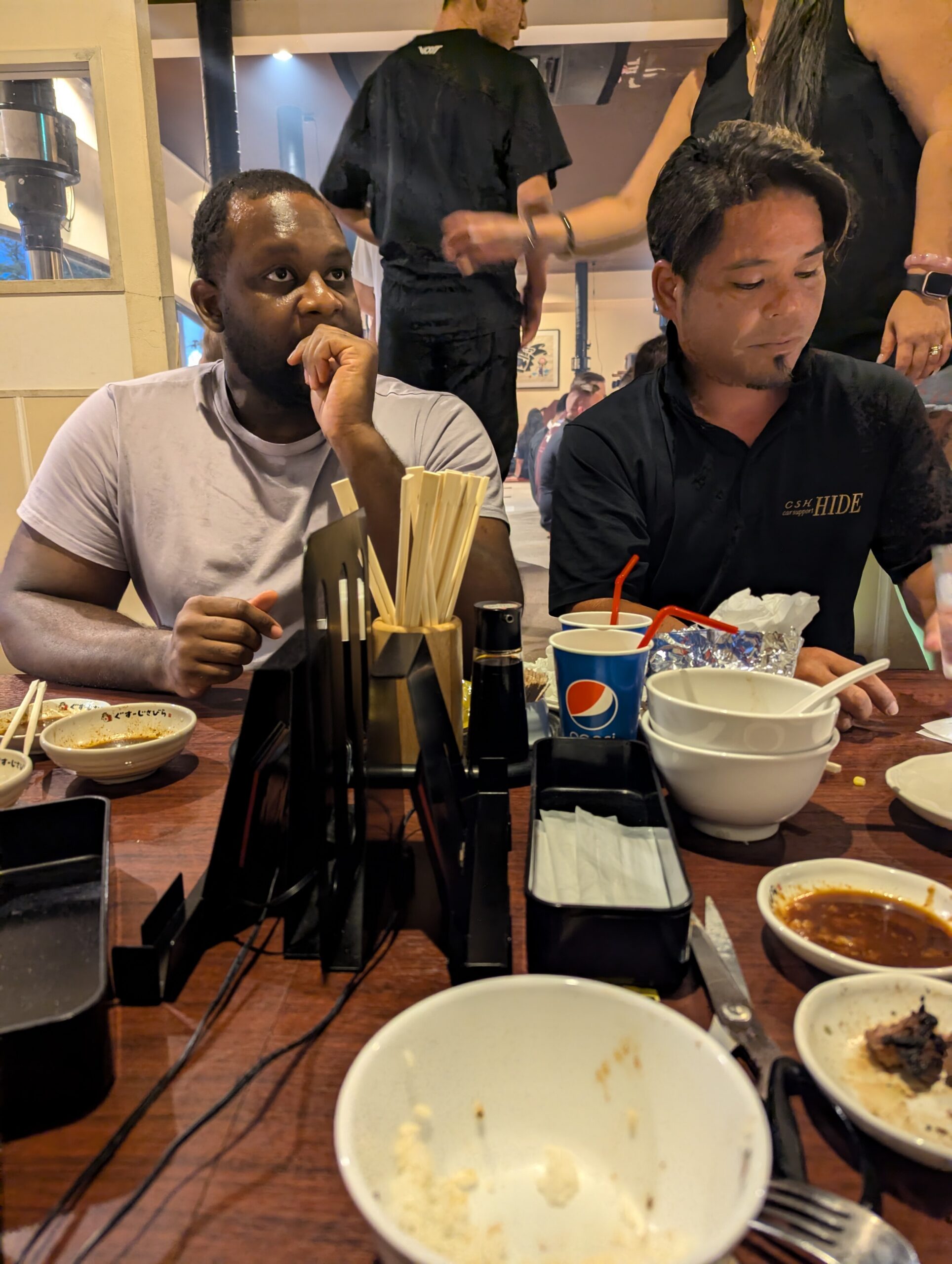 Two men sit at a restaurant table with empty bowls and chopsticks; one looks to the left, the other looks down.