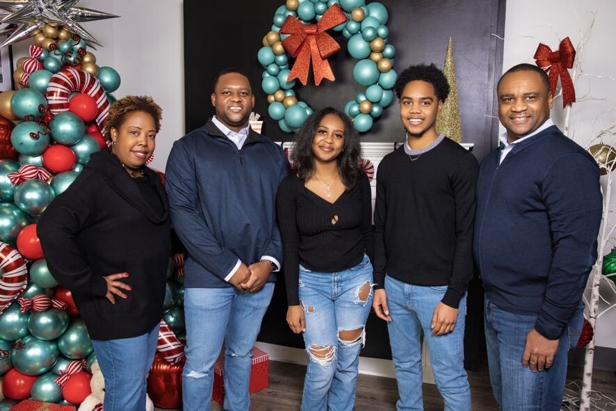 Five adults posing indoors in front of a teal Christmas wreath with a red bow and festive balloon trees; everyone smiles.