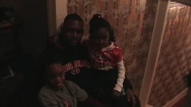 Three people—a man and two children—sit closely together on a couch in a dimly lit room, smiling at the camera.