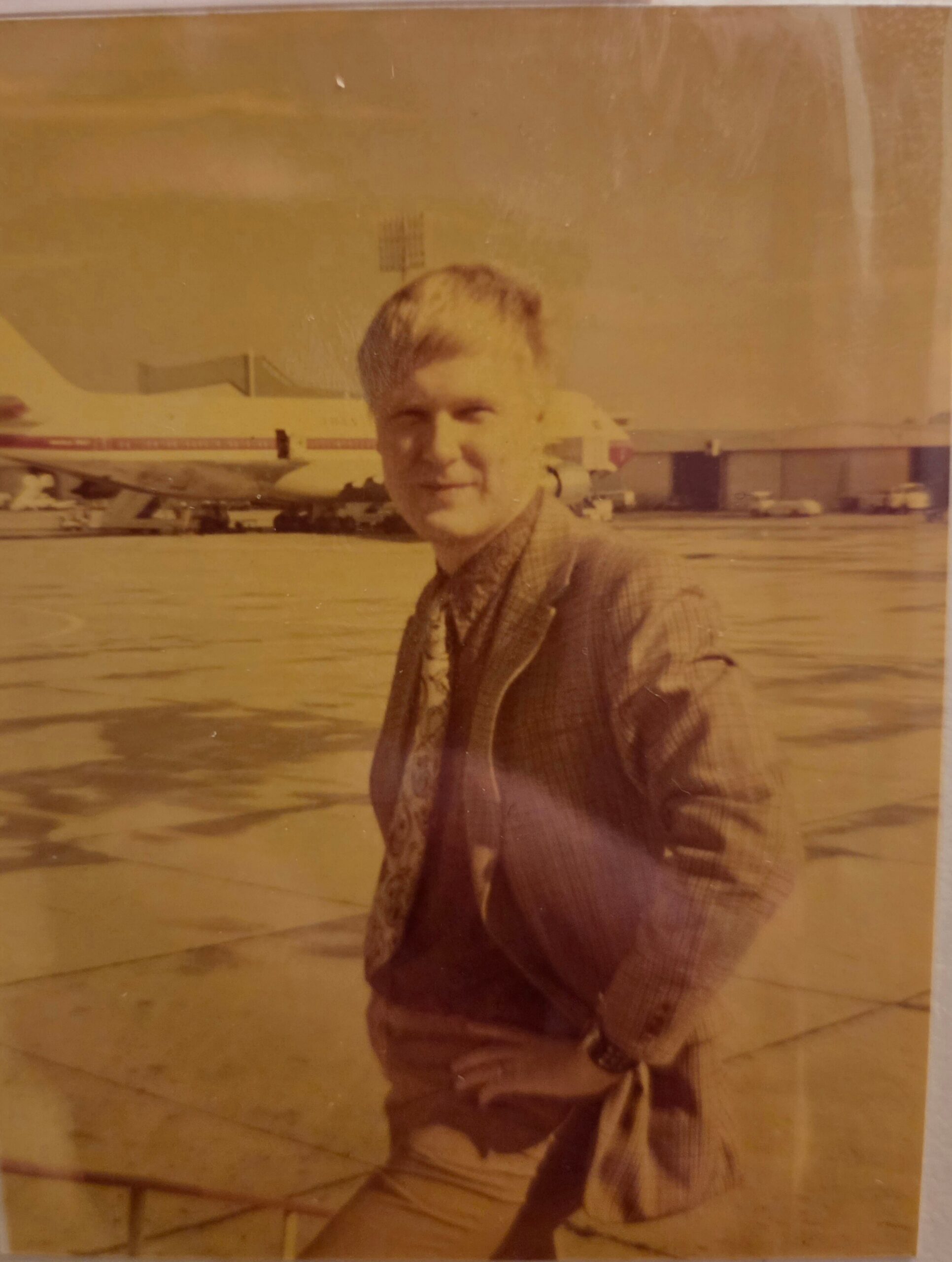 Man in a checked jacket posing with hands on hips at an airport apron, a large white aircraft visible in the background behind him.