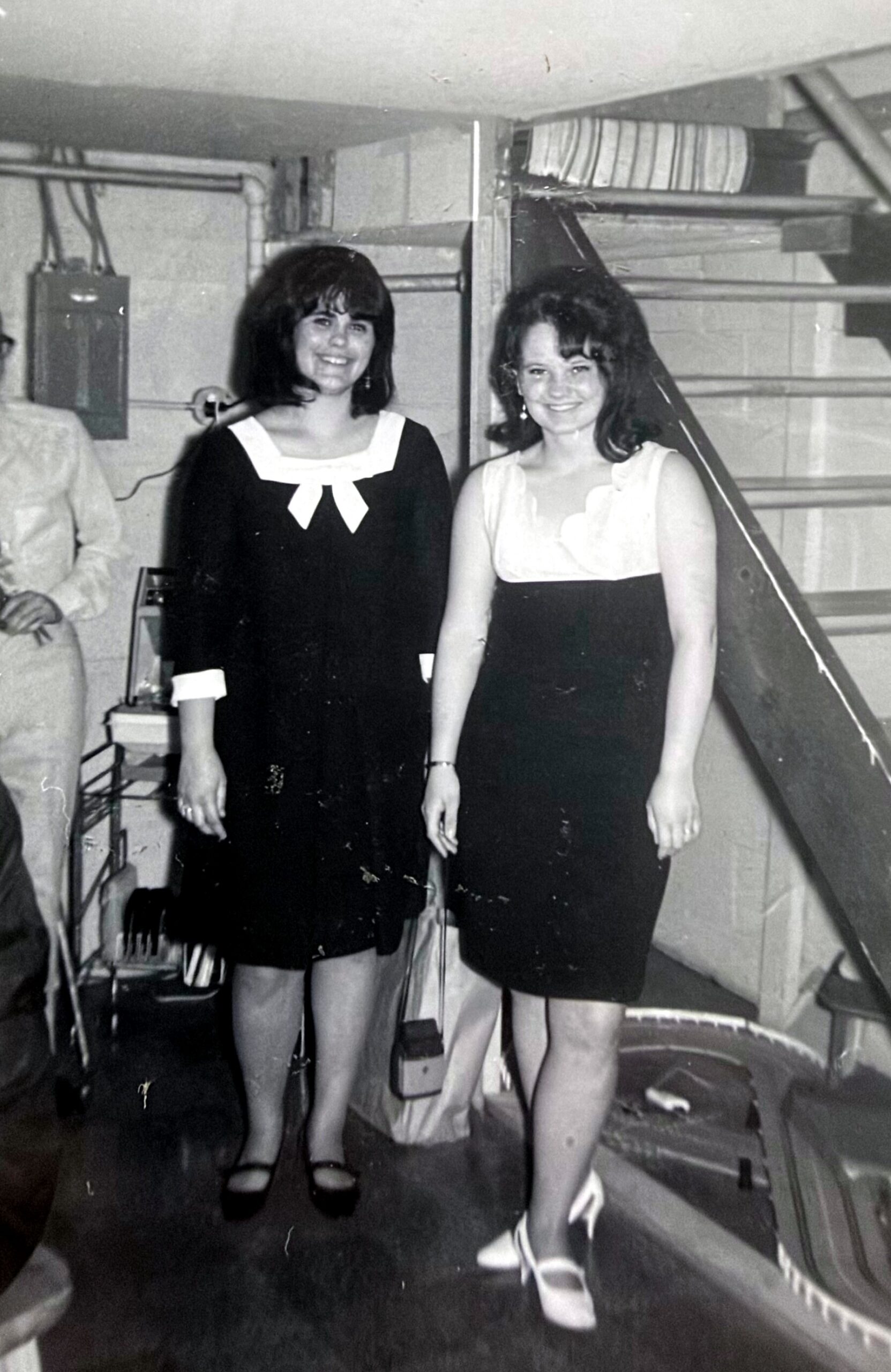 Two women in black‑and‑white formal outfits pose together and smile in a backstage stairwell setting.