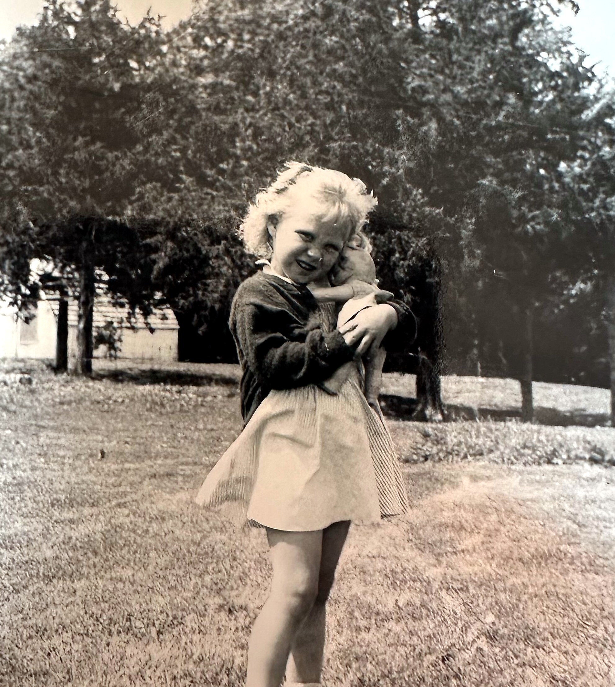 Black-and-white photo of a young girl in a plaid dress outdoors, smiling as she holds a small pet nearby.