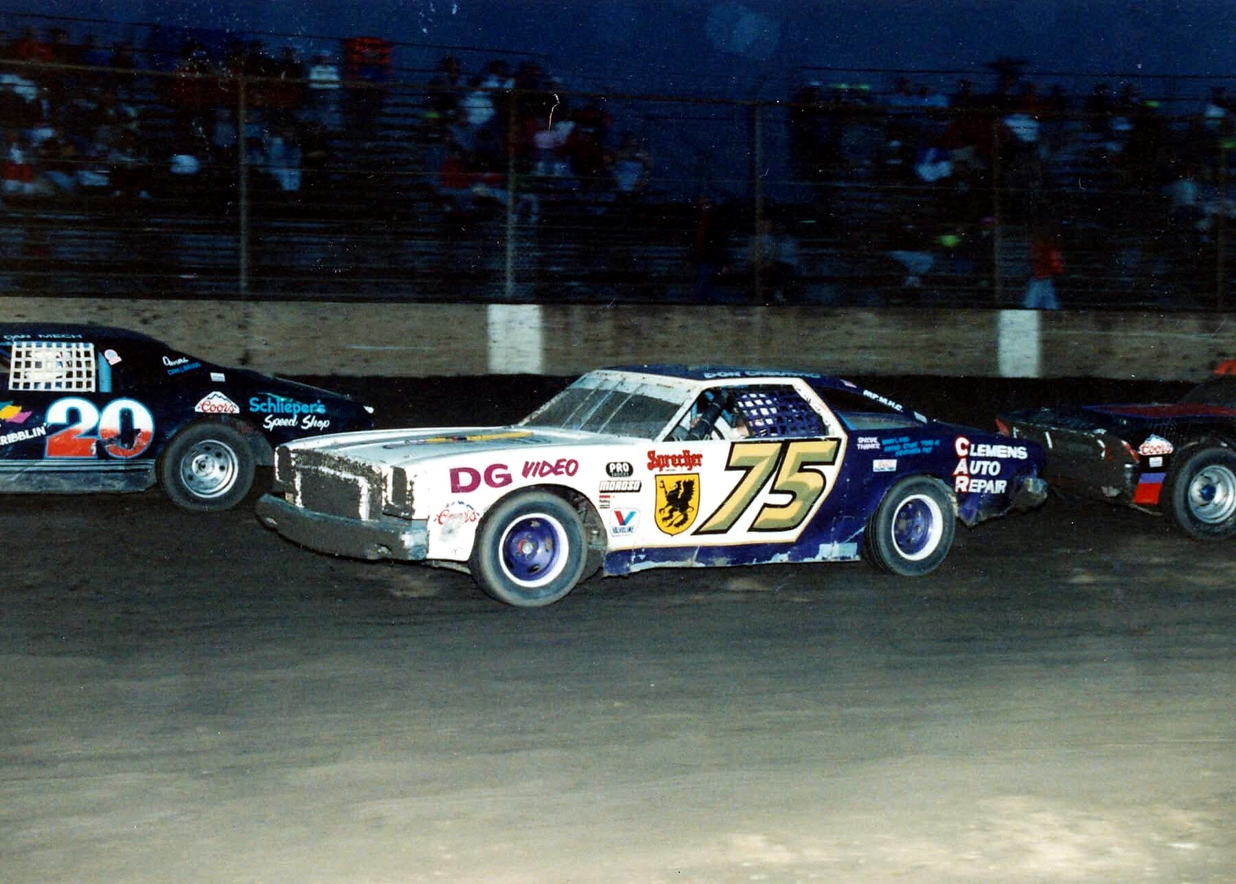 Nighttime dirt-track stock car race with car #75 in the foreground, blue and white with sponsor decals and a cheering grandstand in the background