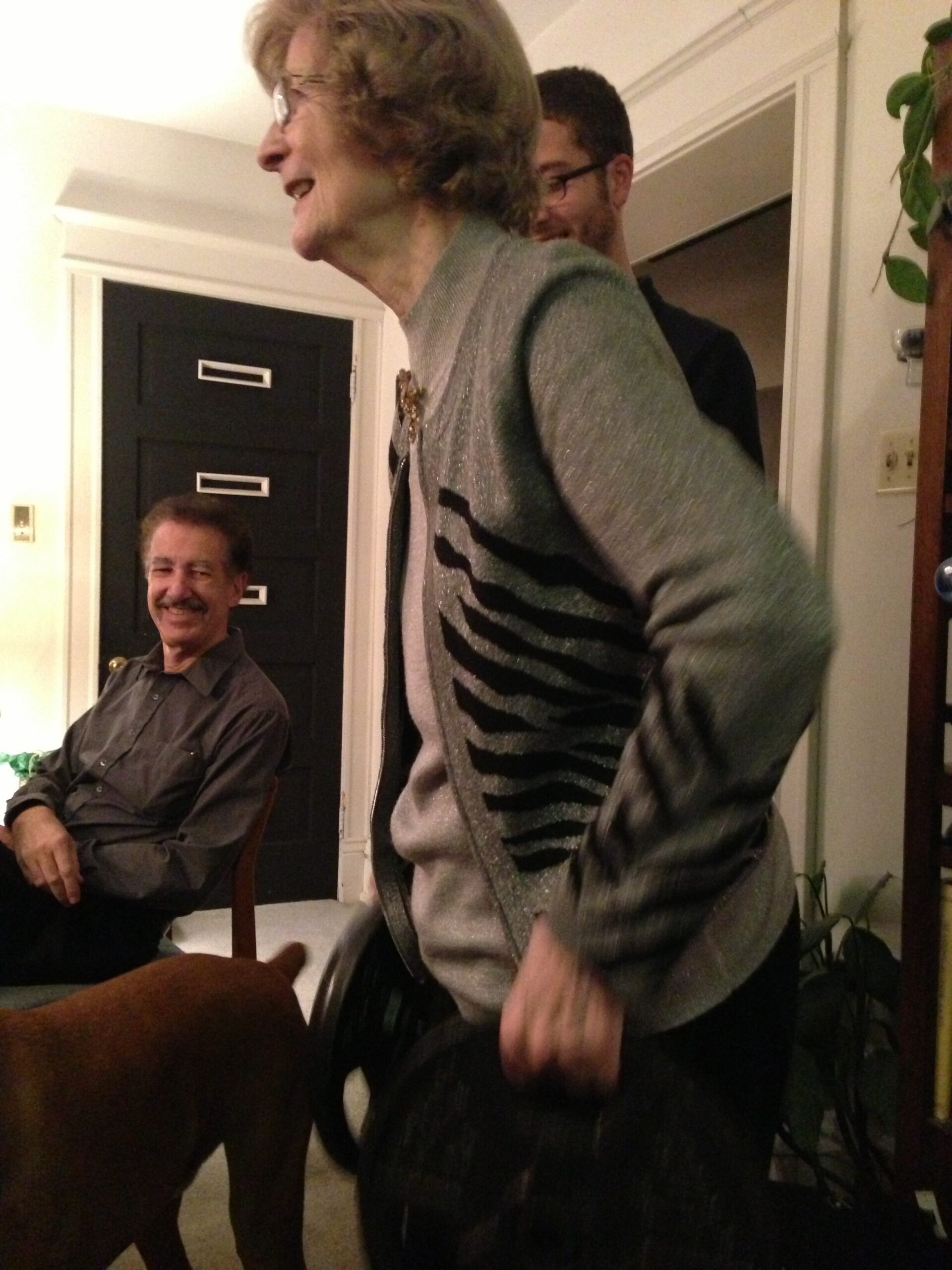 Woman with short gray hair in a sparkling gray cardigan laughing, standing indoors with a man seated in the background.