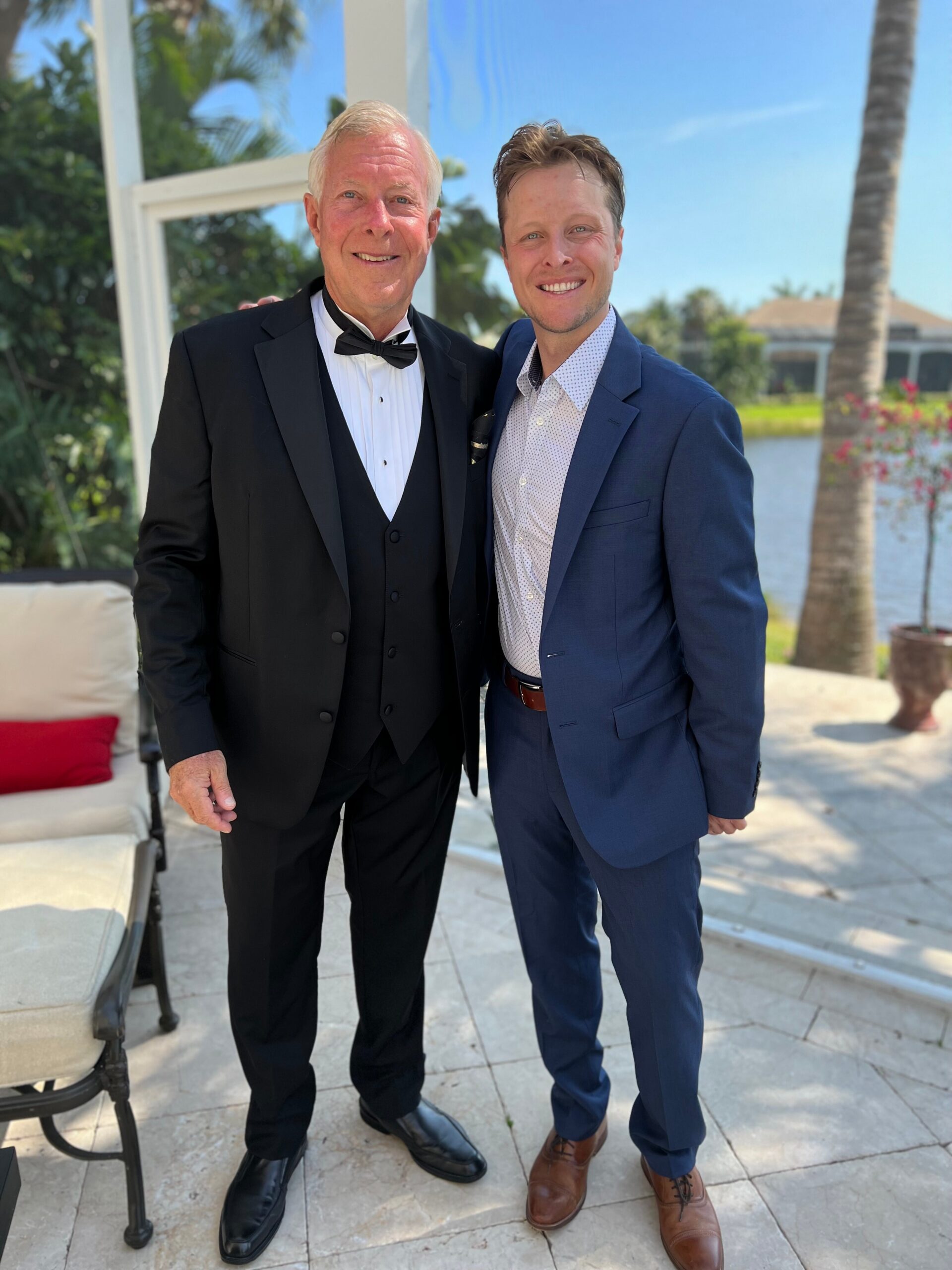 Two men in formal suits posing outdoors, one in a black tuxedo with bow tie and the other in a blue suit, smiling at the camera.