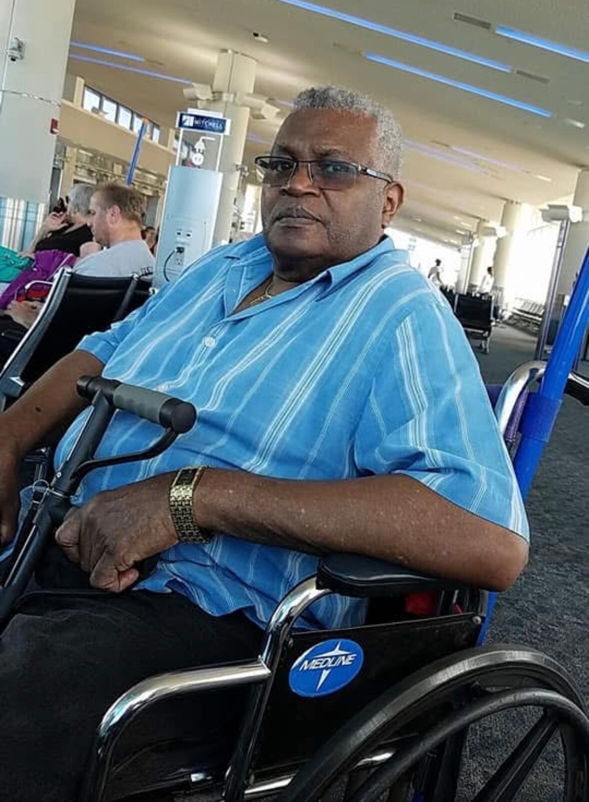Older man with short gray hair and glasses sits in a wheelchair at an airport terminal, wearing a blue striped shirt.