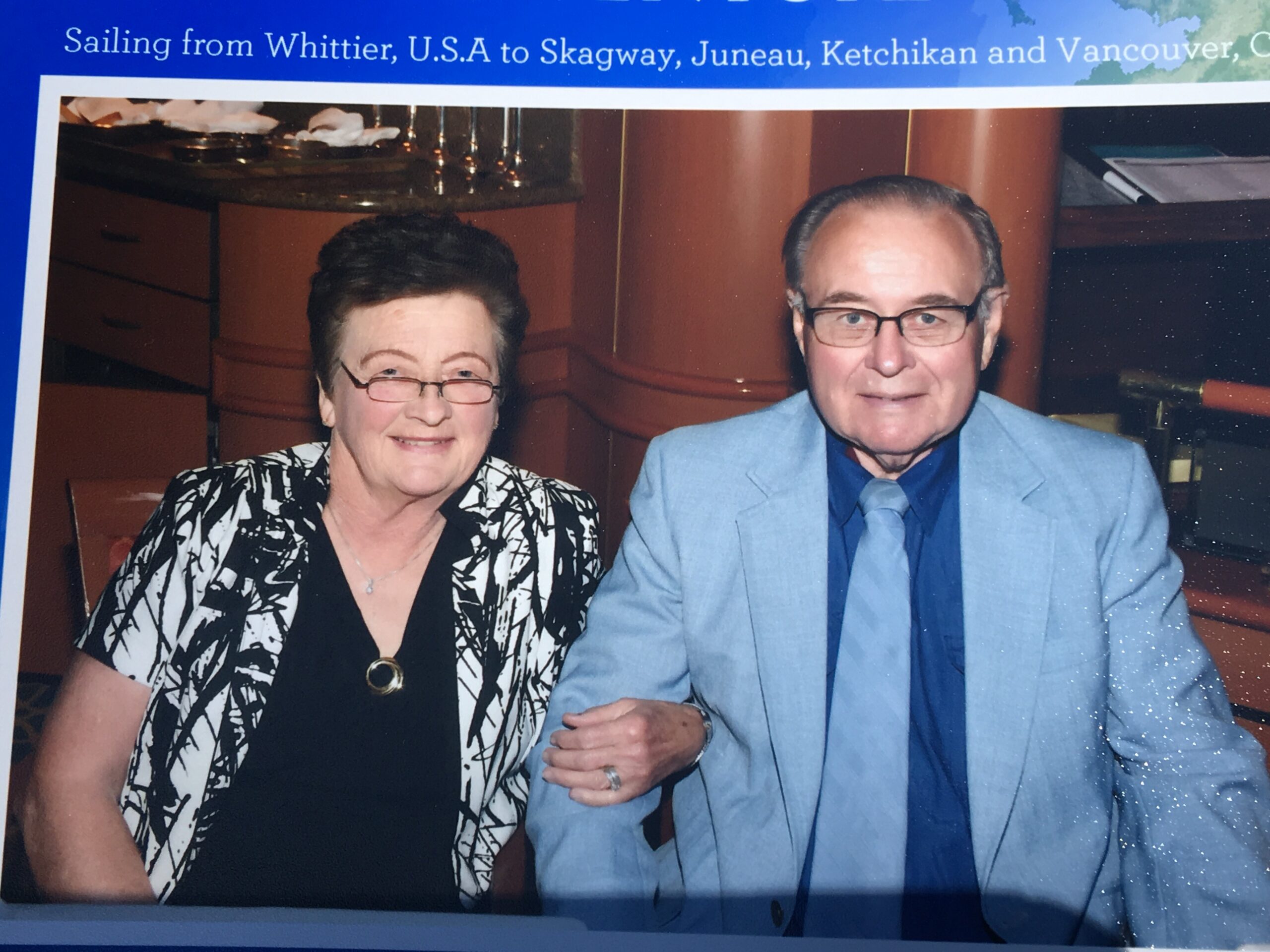 Smiling older couple seated together, woman in a black outfit with a white patterned jacket and the man in a light blue suit and blue tie, their hands joined.
