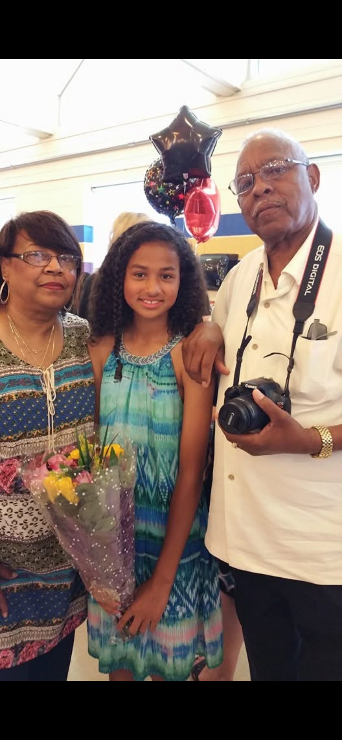 Three generations posing together indoors: a girl in a blue dress stands between a smiling woman holding flowers and an older man with a camera around his neck; balloons float behind them.