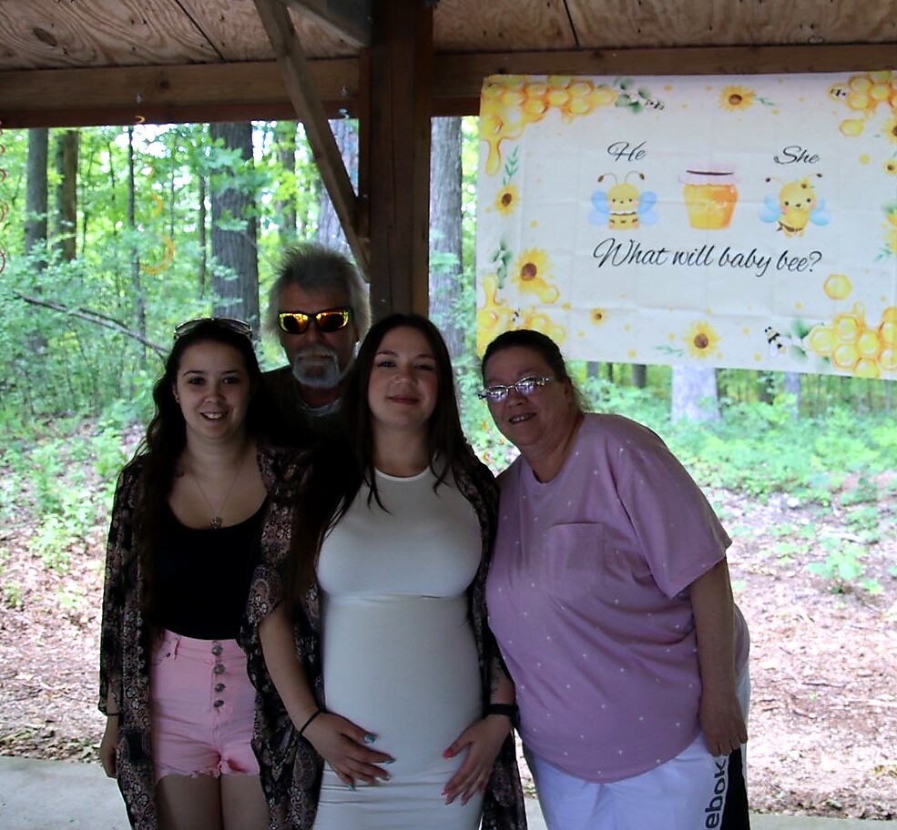 Family of four posing under a wooden shelter at a bee-themed baby shower, pregnant woman in a white dress in the center.