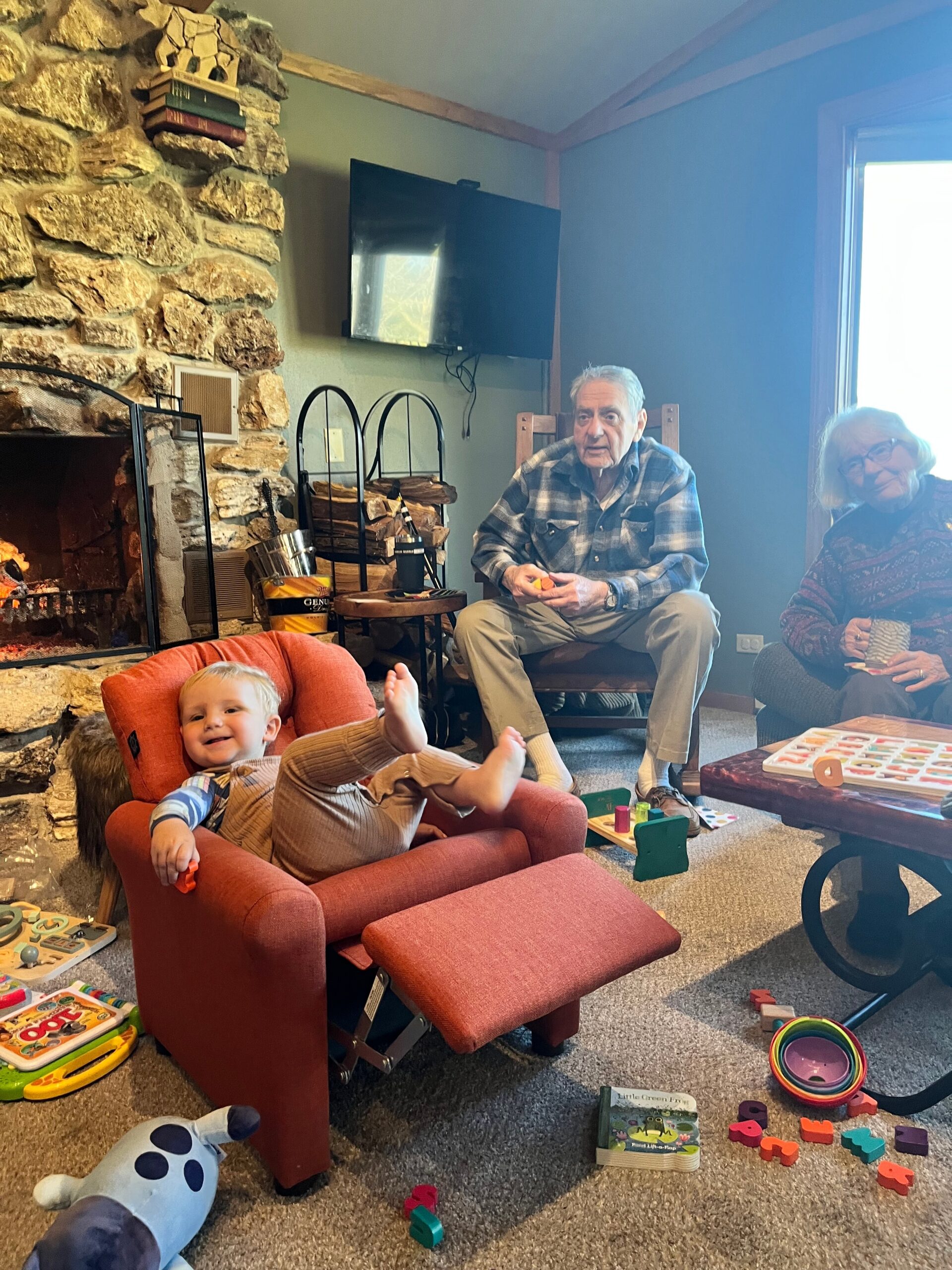 Toddler in a red chair plays in a cozy living room while two seniors sit nearby, with a stone fireplace and toys on the floor.