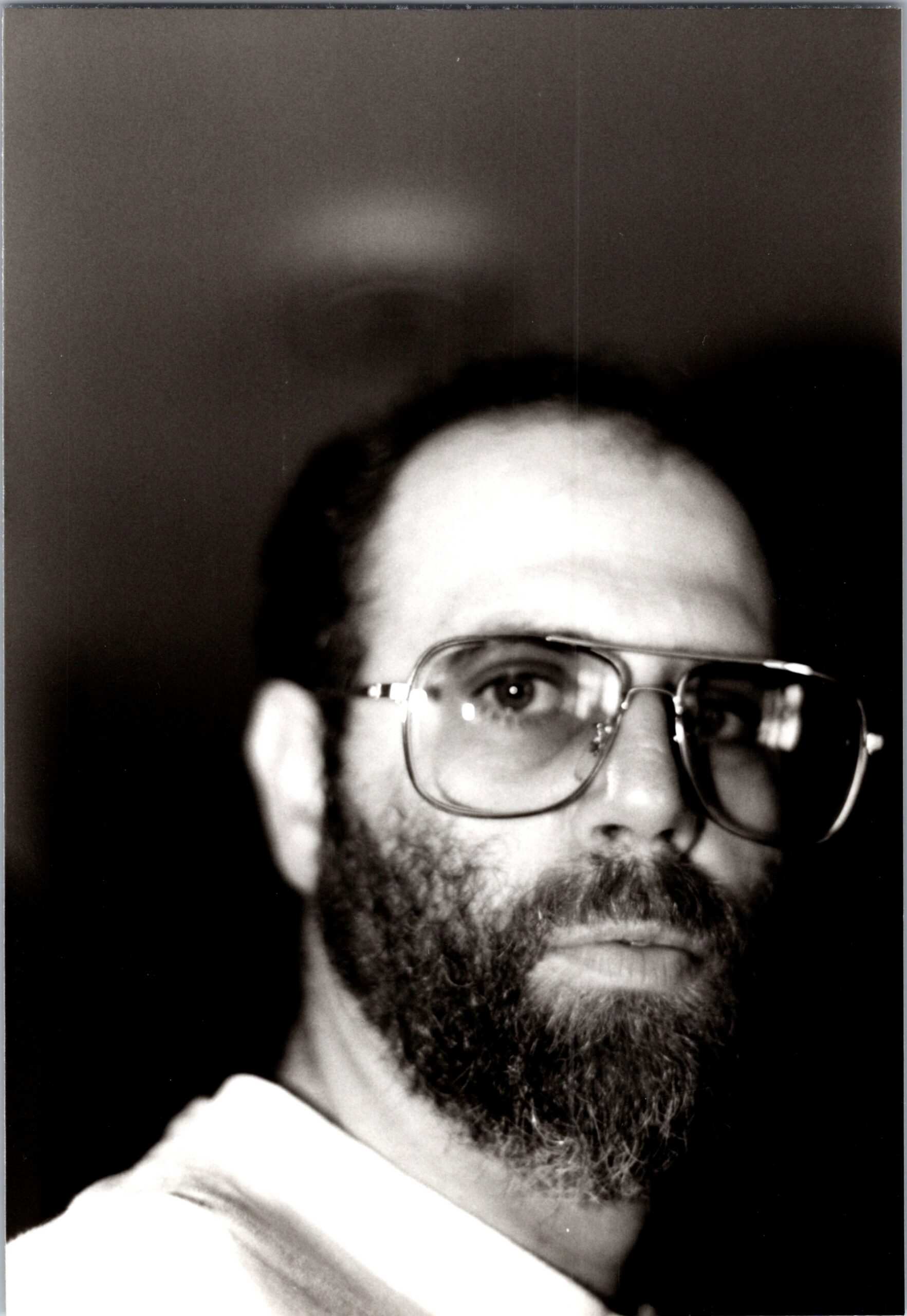 Close-up black-and-white portrait of a man with glasses and a beard, looking slightly upward at the camera.