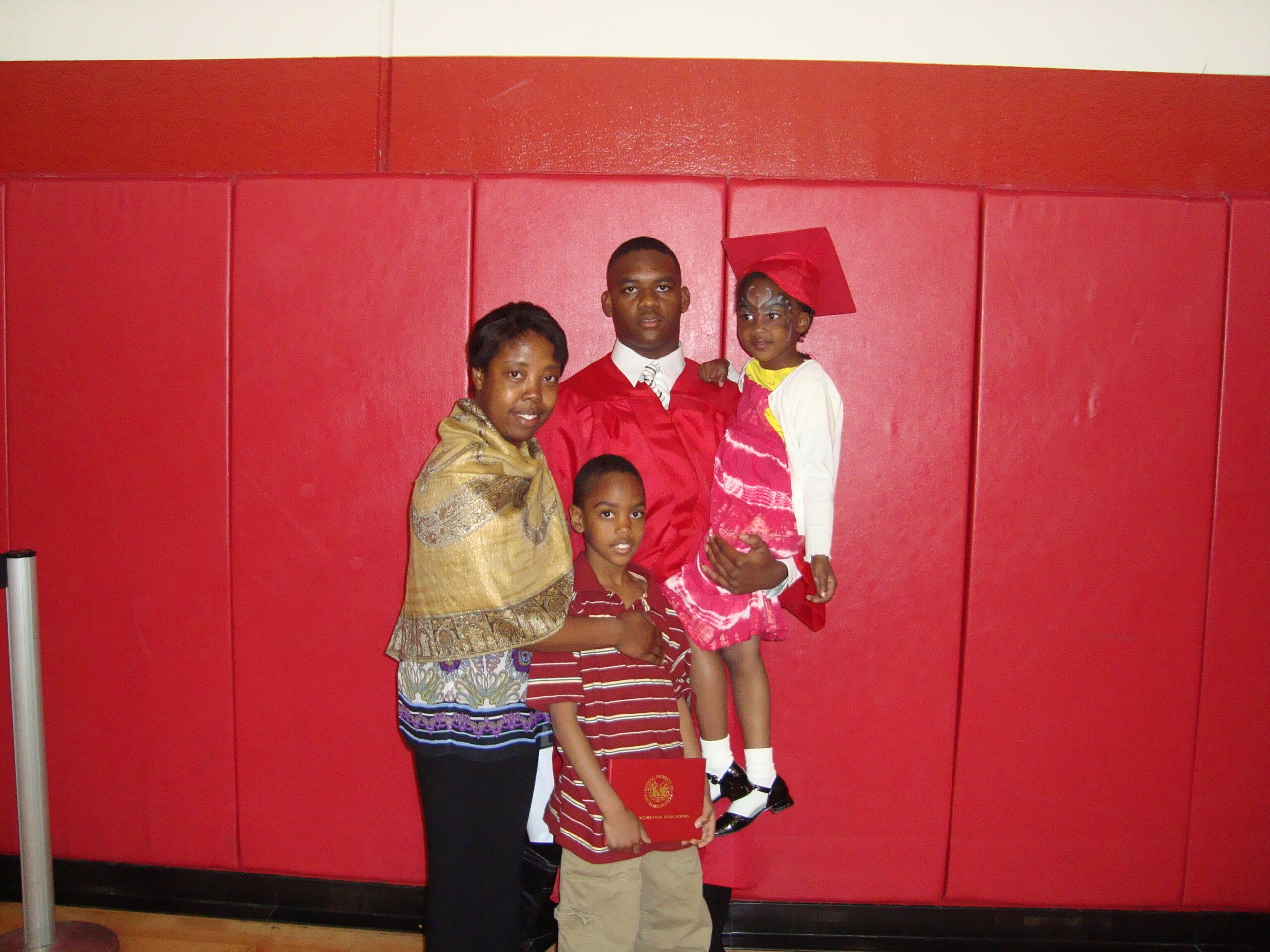Group of five people posing in a gym with red padded walls; a graduate in a red gown holds a red diploma while others smile nearby.