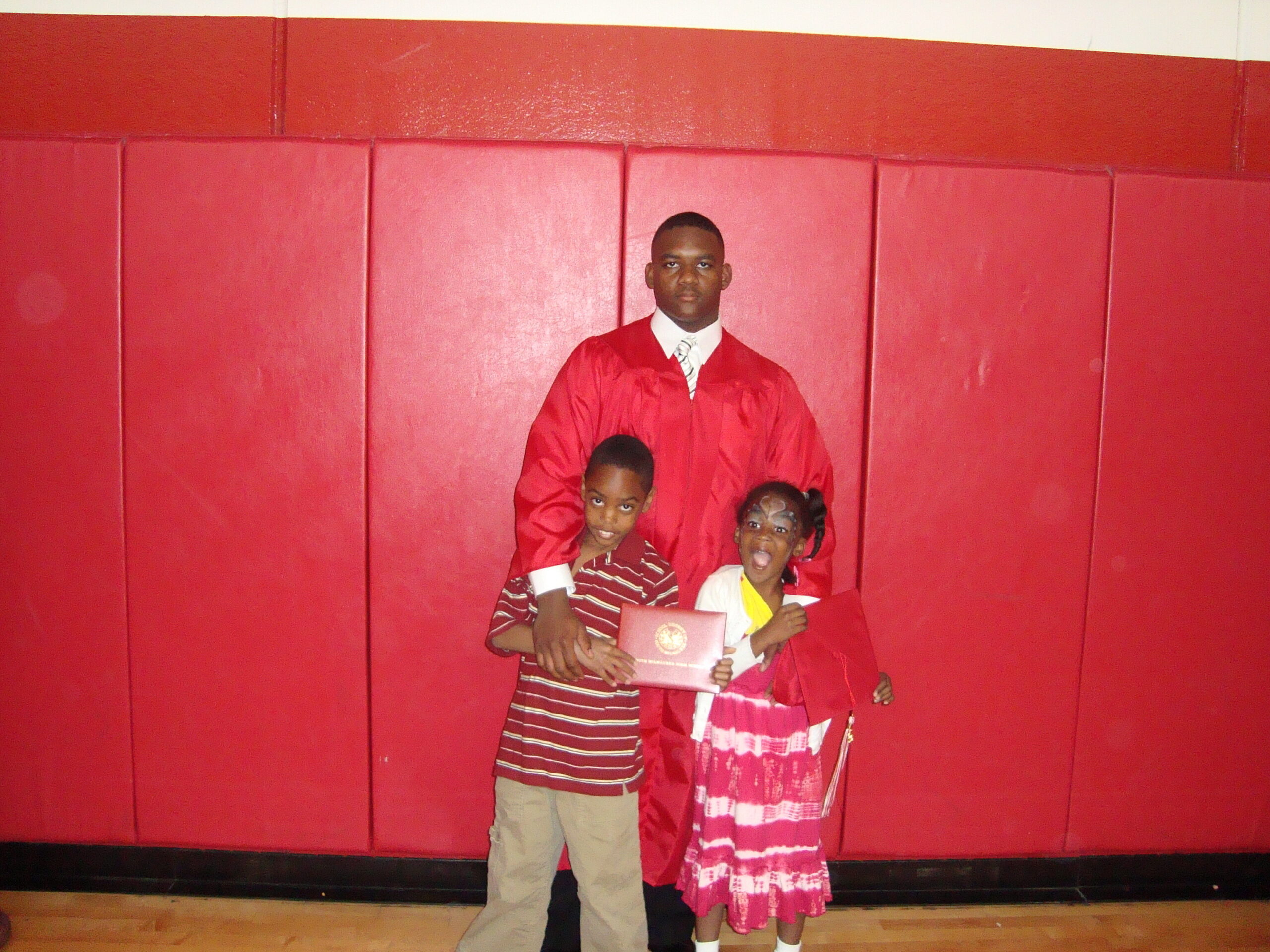 Graduate in a red cap-and-gown stands on a stage with two children holding a diploma, against a red padded wall backdrop.