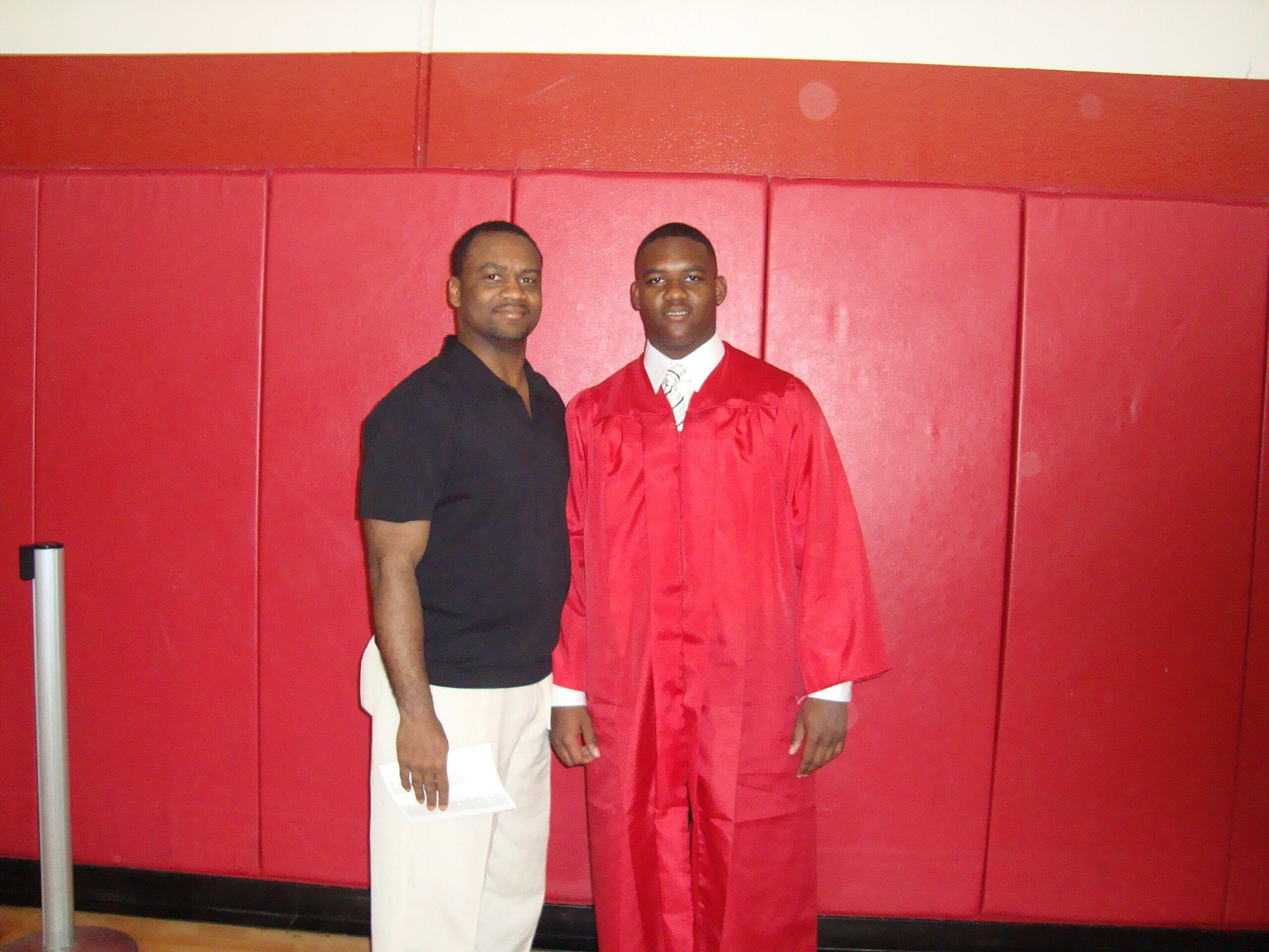 Two men stand side by side; one in a red graduation gown with a white shirt, the other in a black polo, posing against a red padded wall.