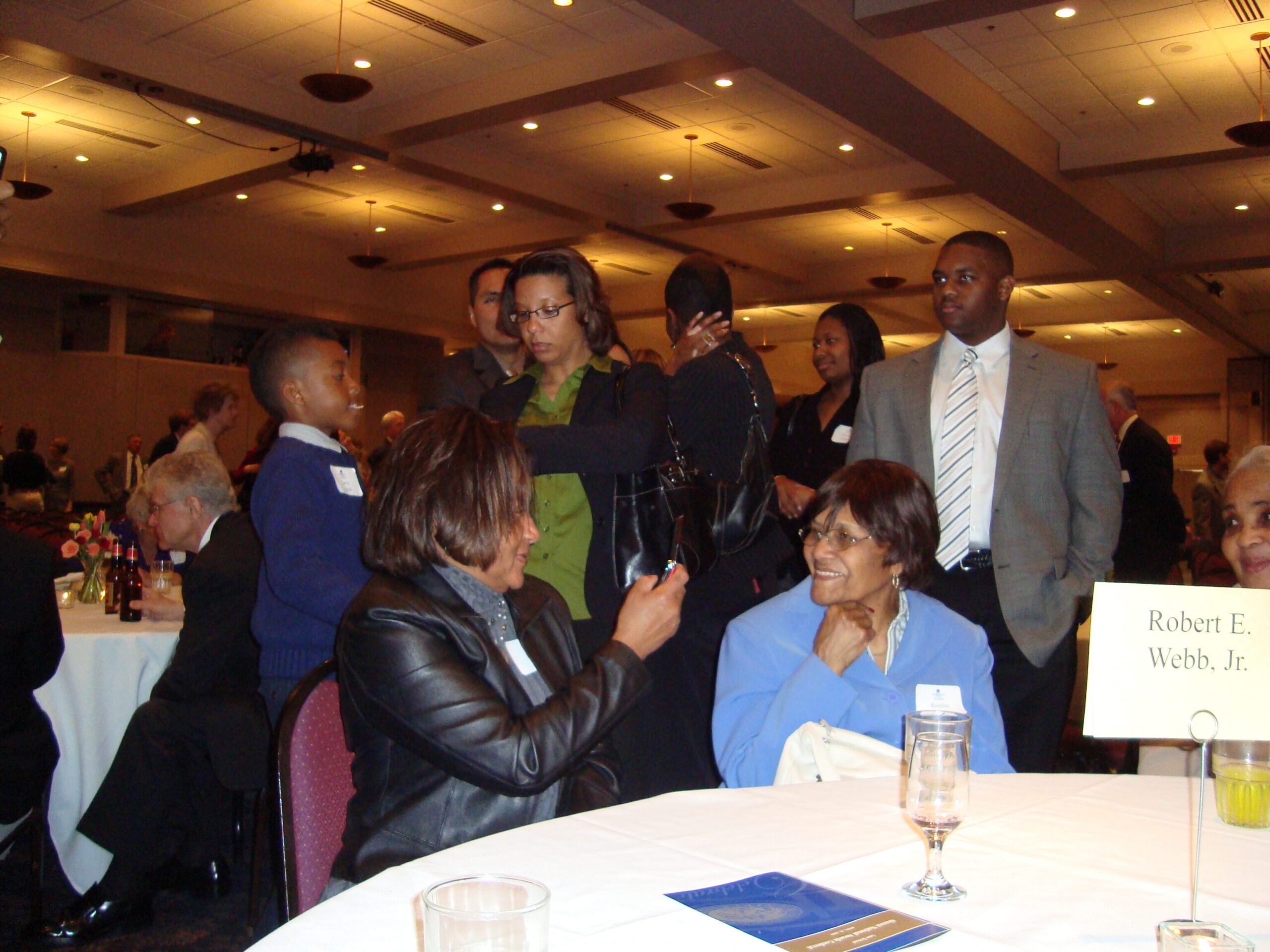 People mingle at a formal dinner in a banquet hall; several stand and chat around tables with name cards visible on a white-clothed table.