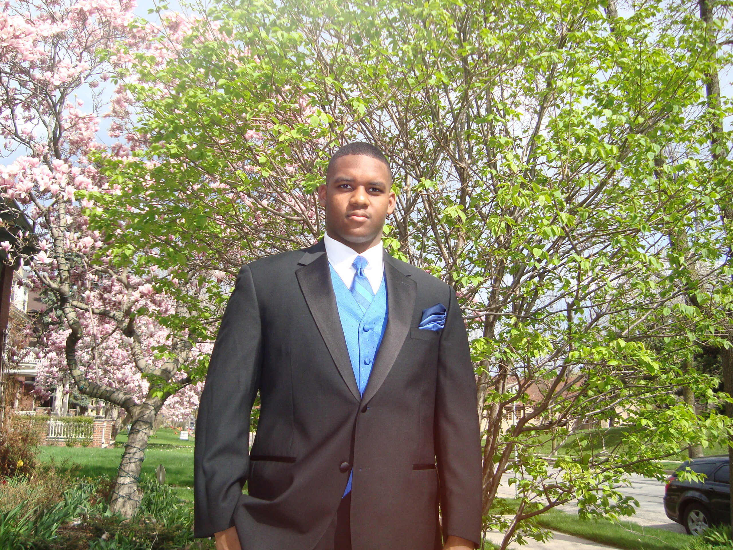 Man in a dark suit with a blue vest and tie posing outdoors in front of blooming pink flowers and green trees in a park setting.