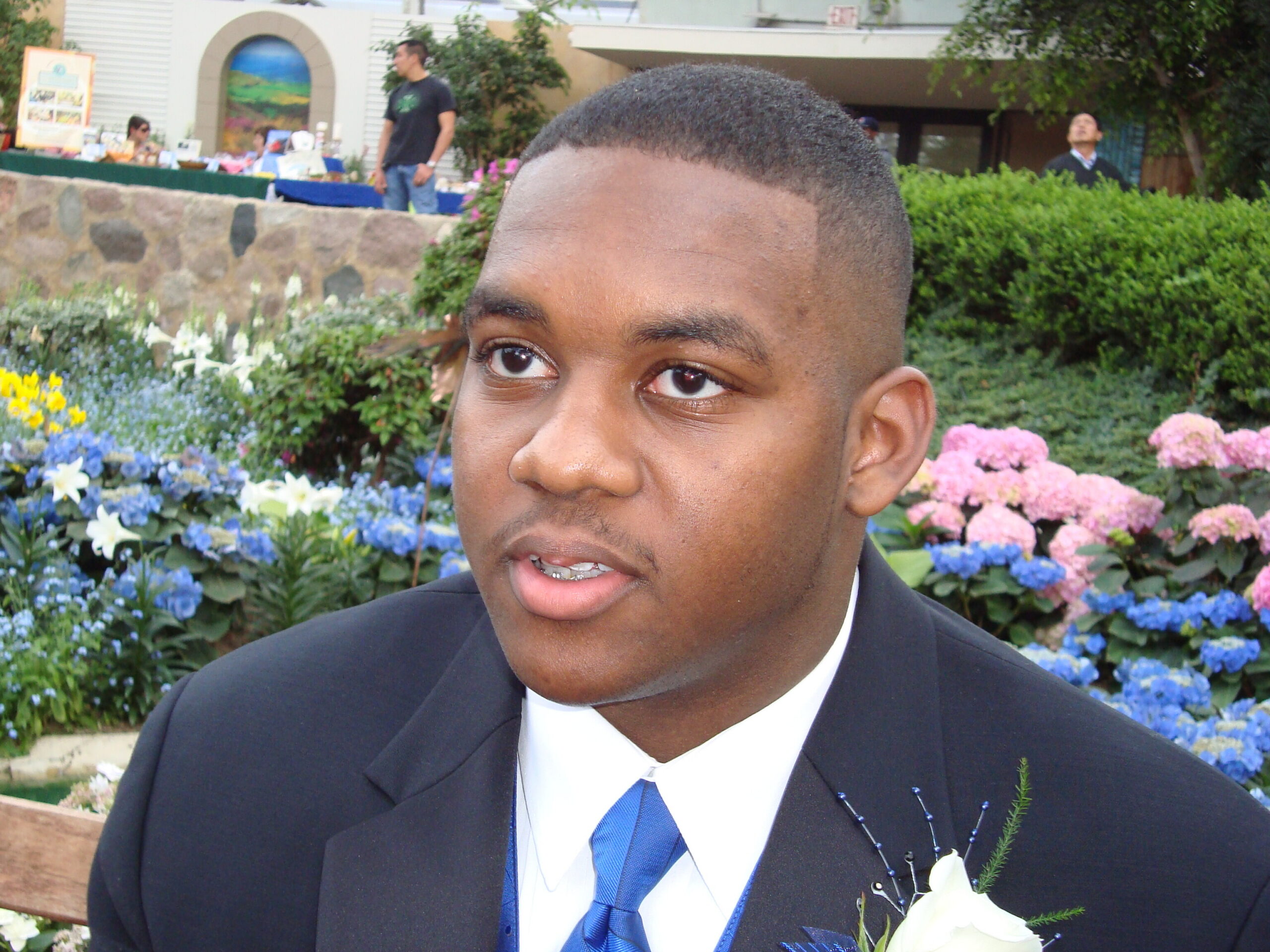 Close-up of a man in a dark suit and blue tie in a garden with colorful flowers in the background.