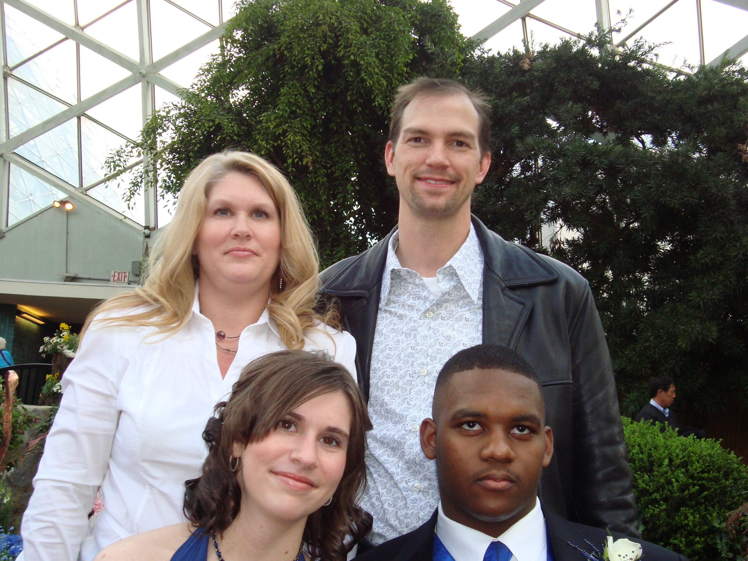 Group of four posing for a photo indoors beside a glass structure and plants: woman in white blouse left, girl in blue dress foreground, tall man in a patterned shirt and leather jacket behind, and a boy in a suit at the bottom right.
