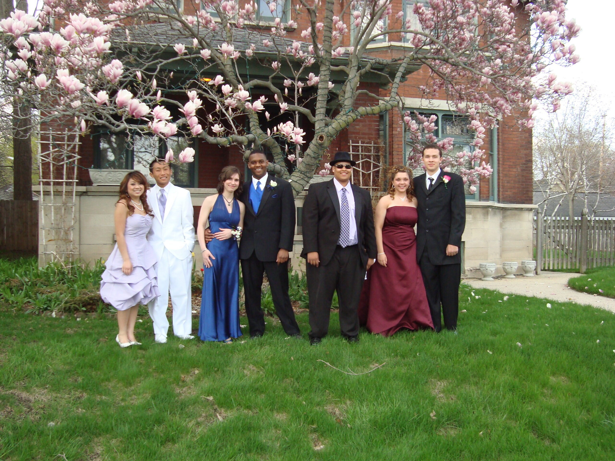 Seven young adults in formal attire pose on a lawn beneath pink magnolia blossoms in front of a brick house.