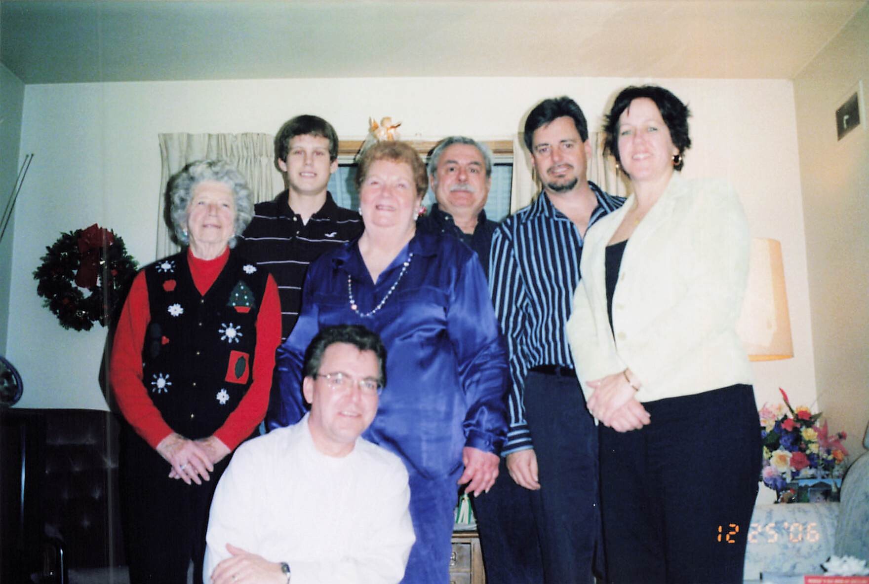 Seven-family group posing for a casual holiday photo in a living room, with a wreath and festive decorations in the background.