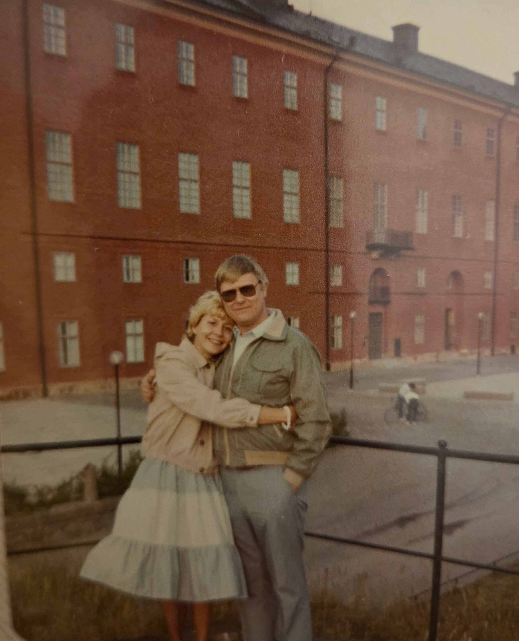 Couple embracing and smiling outdoors in front of a red brick building with many windows behind them, wearing light jackets.