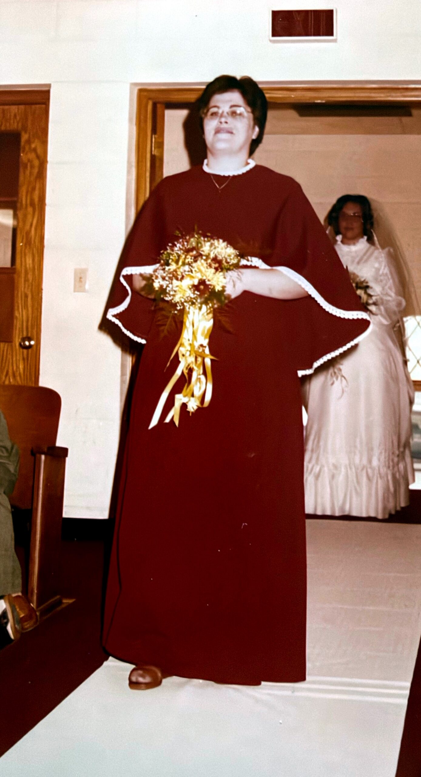 A woman in a burgundy ceremonial robe holding a bouquet at a wedding, with a bride in white in the background, indoors.